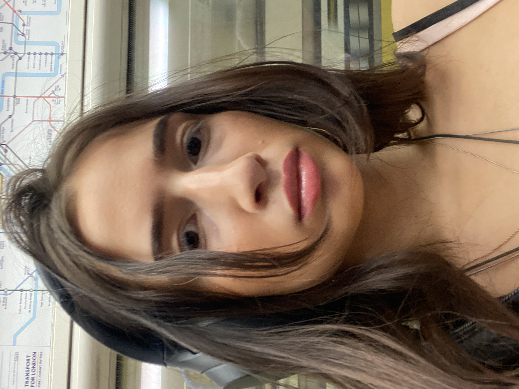 The image is a close-up selfie of a person with brown hair and fair skin, with a London Underground map in the background. The person is wearing lipstick and a thin necklace. The background map has the text "TRANSPORT FOR LONDON" at the bottom. The photo was likely taken on the London Underground train.
