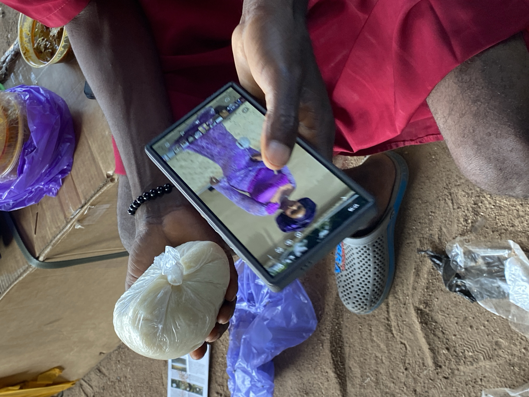 A person's arms and hands are visible, holding a white ball of what appears to be dough or food wrapped in clear plastic. The person is also holding a smartphone, displaying an image of a woman in a purple dress. They are wearing a red garment and a black beaded bracelet. In the background, there is a cardboard box, a purple plastic bag, and a small plastic container with brown food. The ground appears to be sandy and dusty. A grey and blue sandal is also visible on the ground. The scene is likely outdoors, possibly in Jalingo, Nigeria, given the context provided.