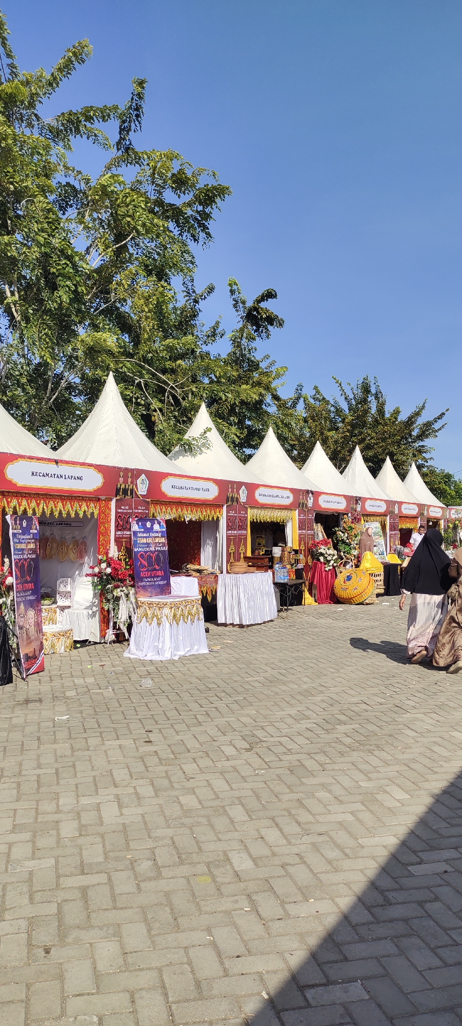 The image depicts an outdoor market scene under a clear blue sky, with trees visible in the background. The foreground is paved with interlocking gray bricks. Several white tents, each with a pointed peak, are set up in a row. Each tent is adorned with red and gold banners. Some banners bear text in Indonesian, including "KECAMATAN LAPANG" and "KECAMATAN TAMPAK PASIR."
Underneath the tents, various stalls are displayed. One stall on the left features two banners with images of people, and the text "ACEH UTARA." There are decorative flowers and white tablecloths covering some surfaces. Further to the right, another stall has a decorative yellow structure and more flowers.
Two individuals are walking away from the camera on the right side of the image. One is wearing a black hijab and a long, light-colored dress, and the other is in a similar long dress. A long shadow is cast across the brick pavement from an unseen object to the right. The overall impression is of a local event or market day.