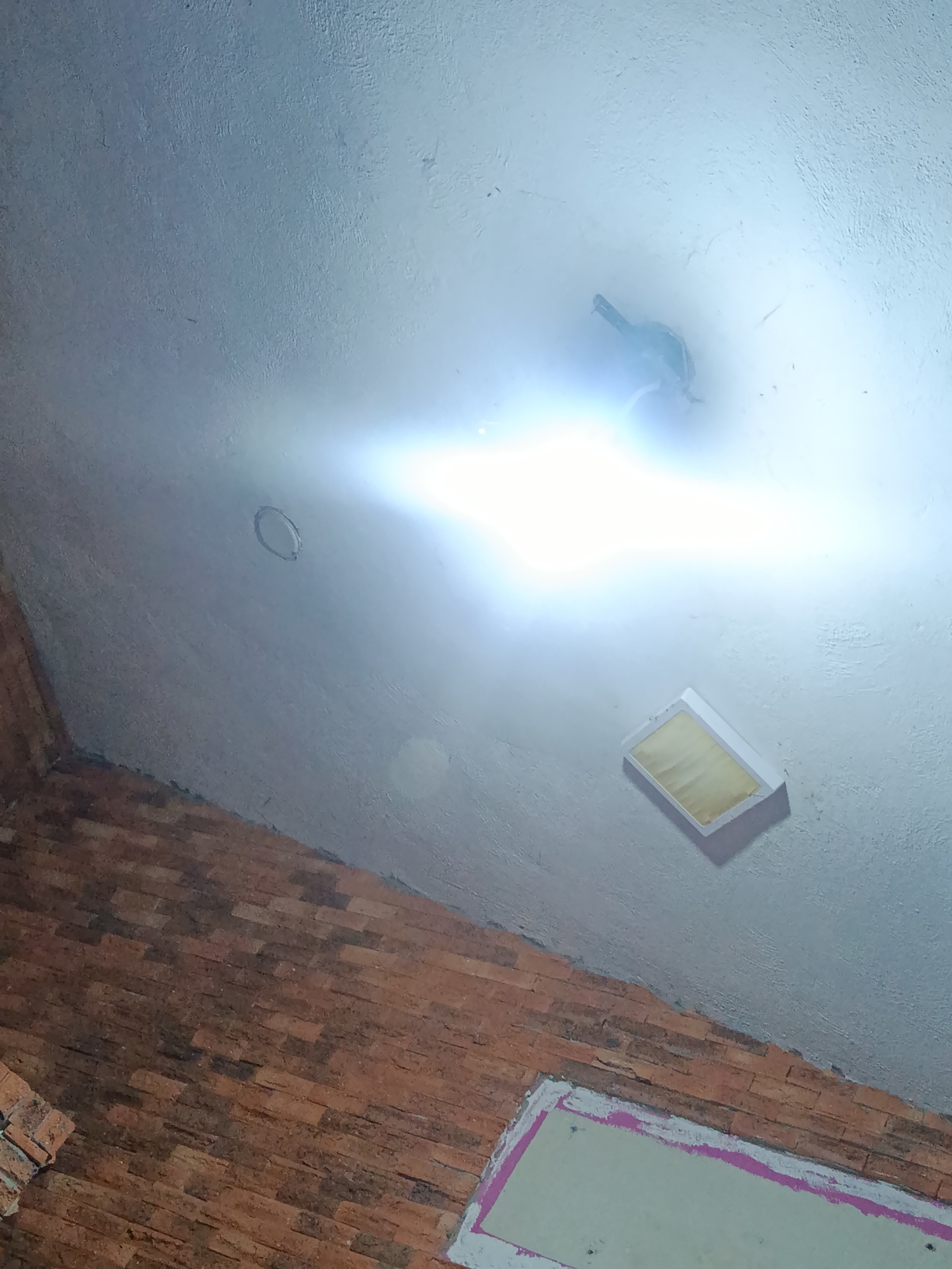 The image shows an interior scene with a partially visible brick wall and a mostly white ceiling. The wall is composed of reddish-brown bricks, and a section of a light green surface with a pink border is seen at the bottom. The ceiling is white and uneven, with a bright, diffuse light source in the upper center. Other light fixtures are present, including a rectangular recessed light near the bottom and a circular opening in the ceiling. The location is unspecified, and no people, subjects, objects, actions, or interactions are identifiable.