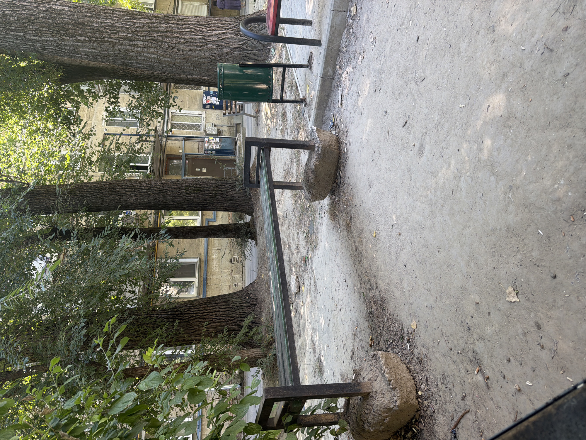 A daytime, eye-level, exterior shot captures a worn, dark green metal park bench with a dark metal frame, supported by rough, concrete blocks that appear to be crumbling. The bench is positioned on a dirt and gravel ground in what looks like a residential courtyard in Chișinău, Moldova. Large, mature trees with rough bark and dense green foliage are prominent in the background, partially obscuring a multi-story apartment building with light-colored walls and white-framed windows. A green metal trash can stands near the bench, and a red-seated bench is partially visible to the right. The ground shows scattered debris, including leaves and cigarette butts. In the distance, a person in a dark dress walks away from the camera. The lighting suggests a sunny day.