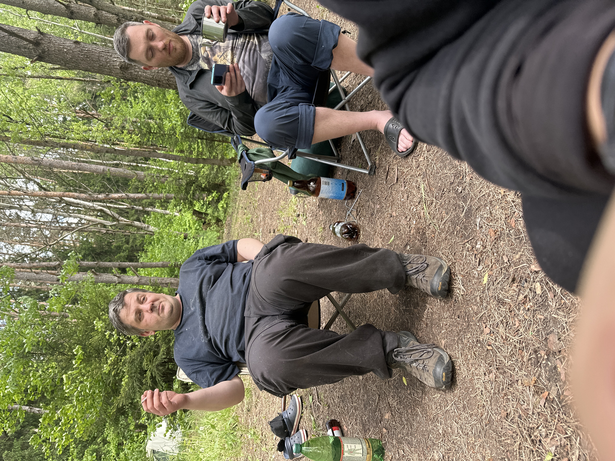 The image depicts a scene in what appears to be a forest clearing in Папярнянскі сельскі Савет, Belarus. Two men are present. One man is sitting in a folding chair, wearing a dark t-shirt, dark trousers, and hiking boots. He is turned towards the camera and is holding a cigarette in his hand. Several empty bottles, including a large green plastic bottle and smaller glass beer bottles, are at his feet. Another man is visible in the background, also sitting in a folding chair. He is wearing shorts, a t-shirt, and a jacket. He is holding a cell phone in one hand and a metal mug in the other. There are trees and green foliage surrounding the clearing. The ground is covered in dry leaves and pine needles.