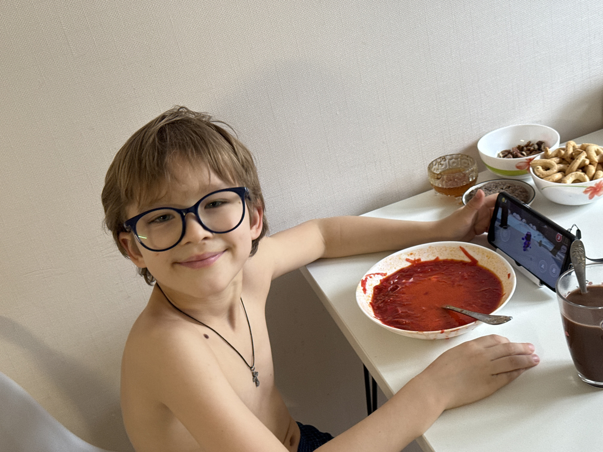 A young boy with light brown hair and distinctive dark-rimmed glasses is captured looking directly at the camera with a gentle smile. He appears to be seated at a white table, and his upper body is bare, revealing a dark necklace with a small pendant. A noticeable mole is present on his right shoulder.
In front of the boy, on the white table, there is a large white bowl filled with a vibrant red, liquidy dish, resembling a soup or stew, with a spoon resting inside it. To his right, he holds a black smartphone horizontally in his hand; the screen is illuminated and displays what appears to be a video game or application with colorful graphics and on-screen controls. The phone also seems to have external grip or controller attachments on its sides.
Adjacent to the smartphone is a clear glass mug containing a dark brown beverage, likely hot chocolate or coffee, also with a spoon inside. Further back on the table, several other food items are arranged: a small clear glass bowl holds a yellowish, viscous substance, possibly honey; another small white bowl contains dark, irregularly shaped items, which could be nuts or dried fruit; and a third white bowl is filled with light brown, ring-shaped snacks, resembling biscuits or crackers.
The background features a plain, light-colored wall with a subtle textured pattern, suggesting an indoor setting. The overall impression is of a casual moment, possibly during the daytime, with the boy enjoying a meal or snack while also engaged with his mobile device.