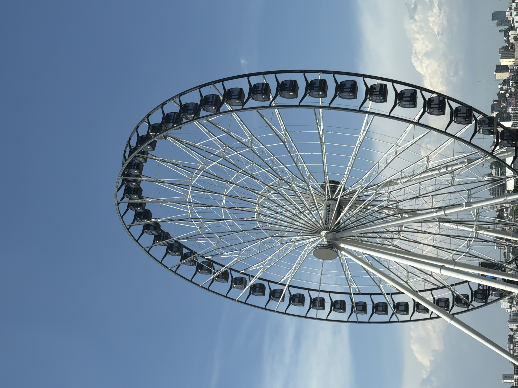A large Ferris wheel dominates the scene, positioned against a clear blue sky with scattered white clouds. The image is oriented with the horizon running vertically, placing the sky on the left and a city skyline on the right. The Ferris wheel is constructed from white metal, featuring numerous radial spokes extending from a central hub to a circular outer rim. Dark-colored passenger cabins, or gondolas, are uniformly spaced along the outer rim.
In the background, a dense urban landscape is visible, characterized by numerous buildings and structures, suggesting an active city environment. No people are visible in the photograph. The scene depicts a bright daytime setting.
Visible text on the central hub of the Ferris wheel includes "GRAN RUEDA", "LA VUELTA", and faintly "CANDELA DELTA", indicating possible branding or a name for the attraction. The overall impression is that of a modern observation wheel in an urban entertainment area.