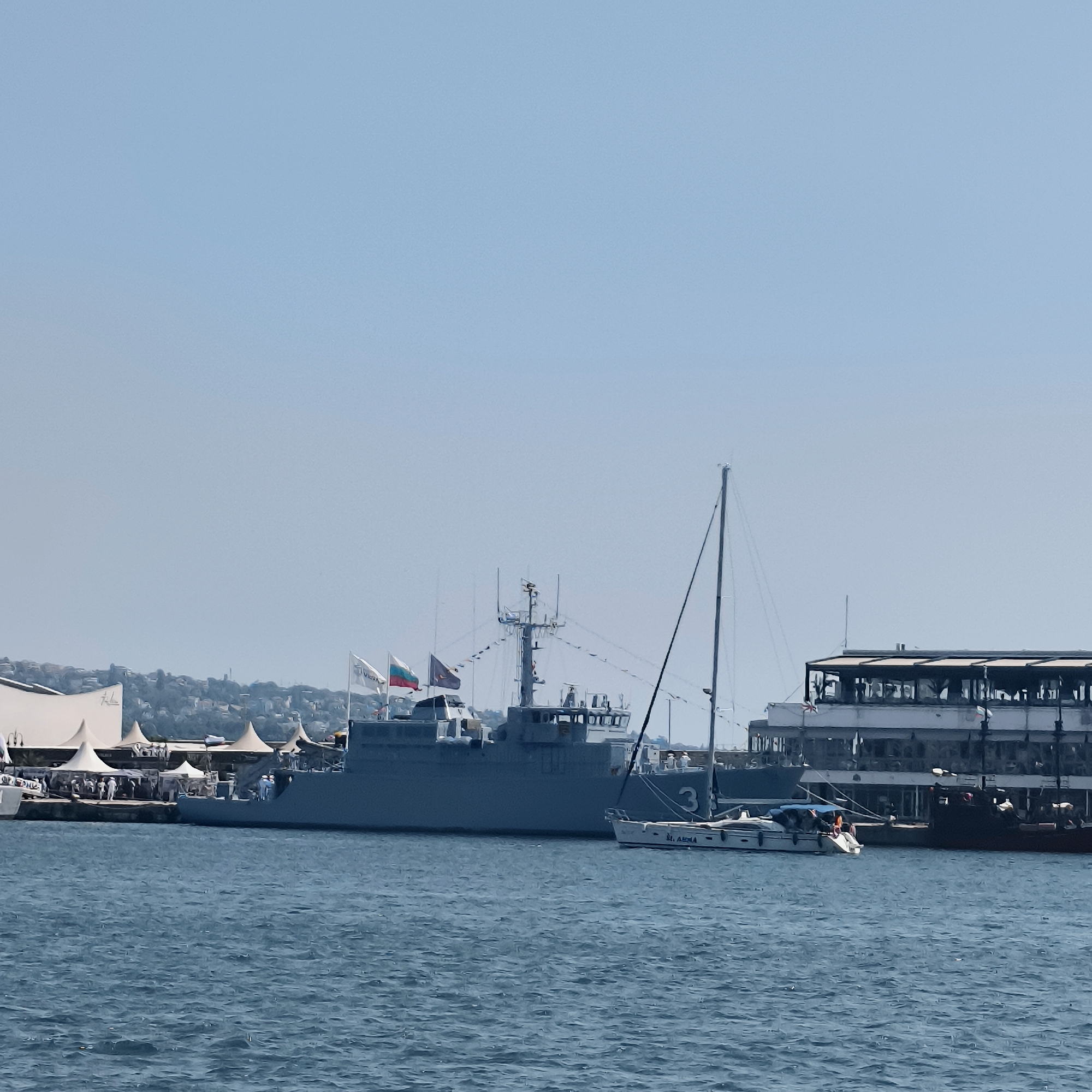 The image depicts a harbor scene in Varna, Bulgaria, with multiple vessels docked and the city's coastline visible in the background. A large, gray naval ship is prominently featured, flying several flags, including what appears to be the Russian flag and another tri-color flag. A smaller white sailboat with the number "3" on its hull is moored to the right of the naval ship. Further to the right, a multi-story building, possibly a restaurant or hotel, is situated along the waterfront. The water is choppy and blue, indicating a sunny day with a clear sky. Several white tents are set up on the dock, suggesting an event or exhibition is taking place. A few people are visible on the deck of the naval ship and near the tents on the dock. The overall atmosphere is one of a busy port with naval presence.