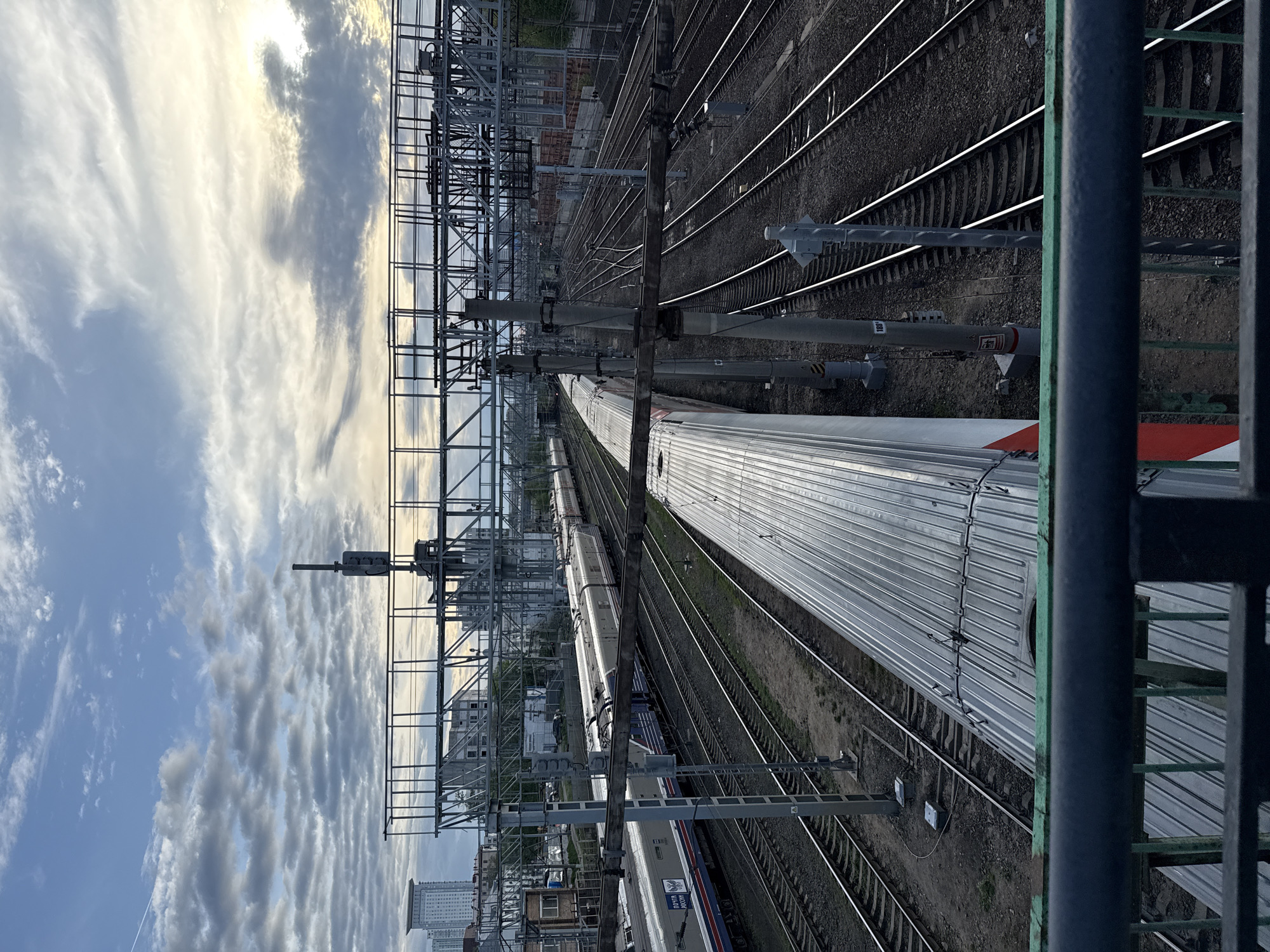 The image, taken from an elevated position, shows a railway yard in Moscow, Russia. Multiple tracks run parallel to each other, occupied by several trains. The nearest train is silver with a red and white stripe at the end. Overhead electrical infrastructure, including wires and support structures, is visible above the tracks. In the background, buildings of varying heights dot the cityscape. The sky is partly cloudy.