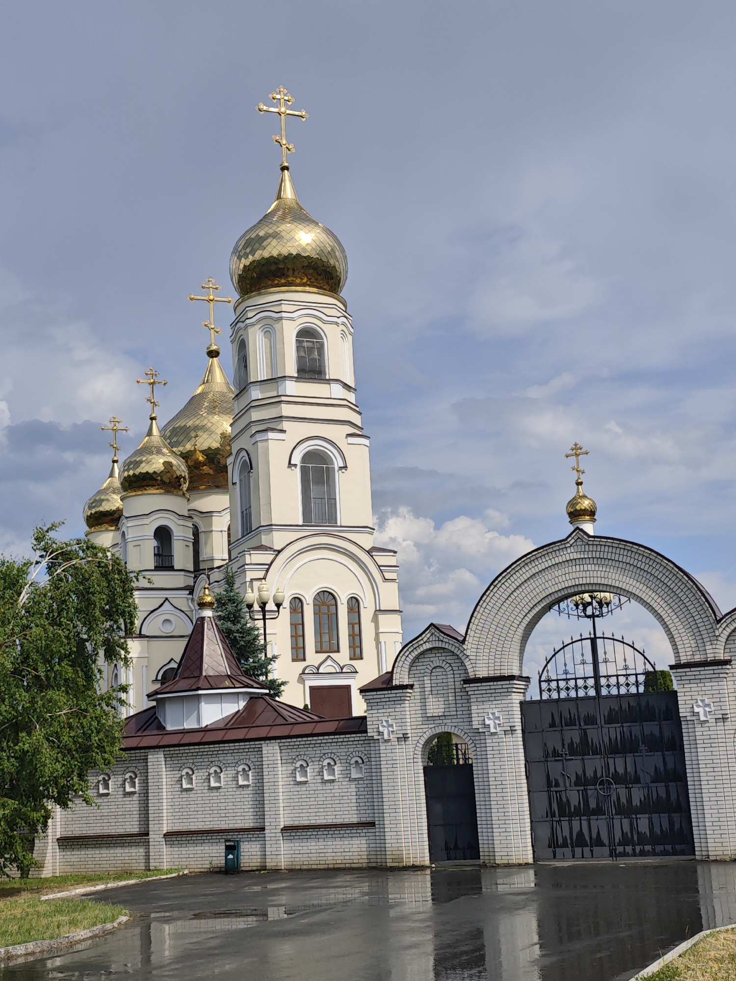 An exterior, eye-level, medium shot captures the Alekseyevka cathedral in Alekseyevka, Russia, under a cloudy sky. The cathedral features multiple golden domes topped with crosses, and a tall bell tower with arched windows. A white brick wall with decorative crosses and arched openings surrounds the cathedral grounds, leading to a large black metal gate. A wet asphalt driveway in the foreground reflects the sky and the cathedral. Green trees frame the left side of the image. The scene appears peaceful and serene.