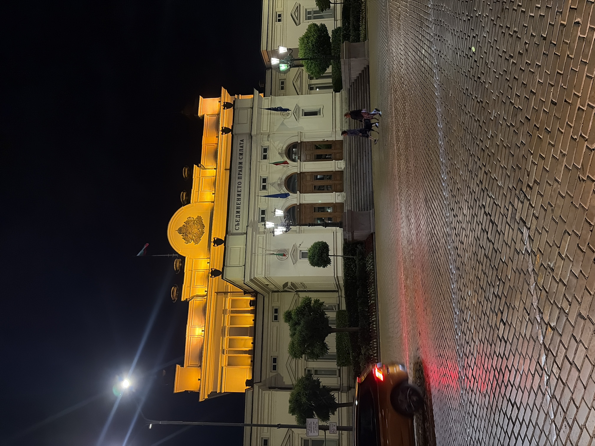 The image depicts the National Assembly Building in Sofia, Bulgaria, at night. The building's facade is illuminated by warm yellow lights, highlighting its classical architectural features, including columns and a prominent inscription above the main entrance. A Bulgarian flag is visible flying from a flagpole.
In the foreground, a cobblestone pavement reflects the lights. An orange car is captured in motion on the left side of the frame, its taillights emitting red light. Two individuals are walking their dog on the central plaza in front of the building. Street lamps with traditional designs are also present. The sky is dark, indicating nighttime. The scene suggests a public space in the heart of Sofia, with the National Assembly Building serving as the primary focal point.