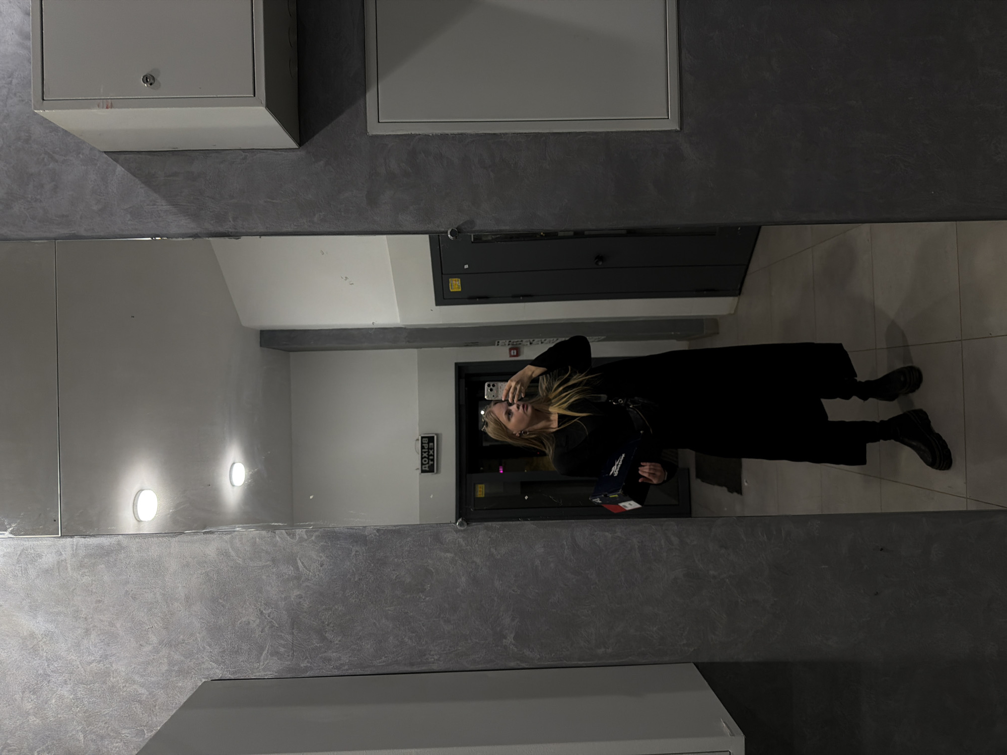 A woman is seen in a full-length mirror selfie. She is standing in what appears to be an indoor public area with tile flooring and gray textured walls. The woman is wearing all black clothing and black shoes and is holding a phone in one hand and a shoebox in the other. Reflected in the mirror are doors and exit signs, as well as lighting fixtures. The location is not identifiable as Izhevsk, Russia, based on the visual information provided.