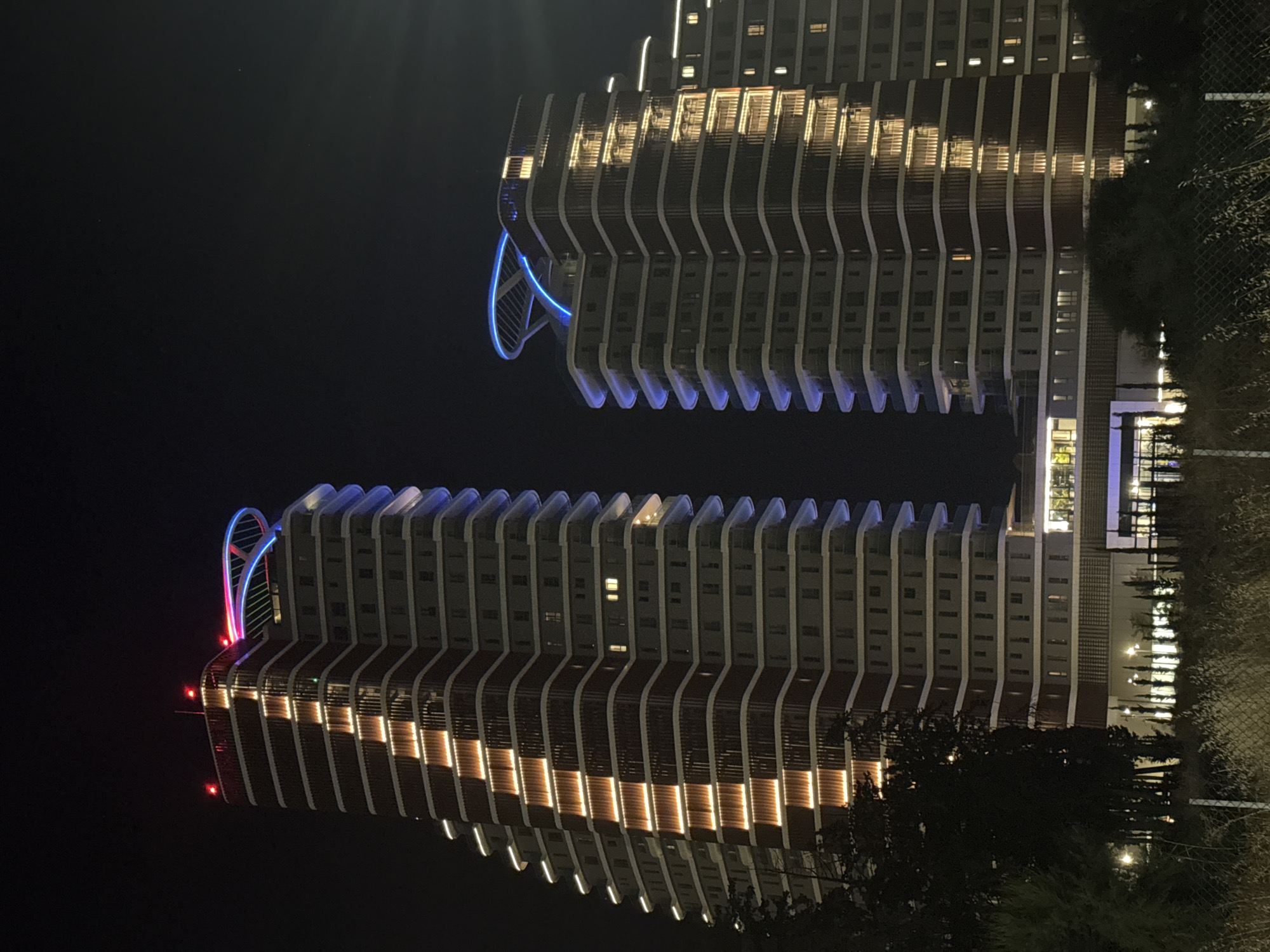 The image depicts two modern, high-rise buildings illuminated at night in the Germasogeia Municipality, Cyprus. The buildings are characterized by their tiered, curved facades and extensive exterior lighting. The building on the left features a prominent vertical strip of warm, orange-yellow lights running up its center, contrasting with the blue and white lights outlining the edges of each floor. The building on the right displays a similar pattern of orange-yellow lighting along its facade, with blue lighting accentuating the upper floors and a distinctive curved blue structure at its apex. Both structures appear to be residential or hotel complexes. In the foreground, there is dense vegetation, including trees and shrubs, and a wire fence is visible, suggesting the photo was taken from ground level across a property line. The sky is dark, indicating nighttime. There are no people or animals visible in the frame.
