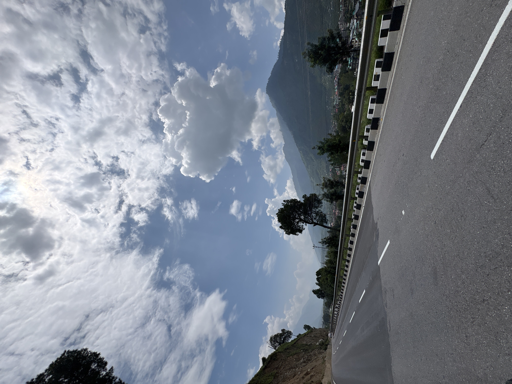 The image depicts a paved road winding through a mountainous landscape. The road surface is asphalt, with white lane markings. A metal guardrail with black and white reflective markers lines the right side of the road. The left side of the road features a steep, rocky embankment.

In the background, rolling green hills and mountains dominate the scene. A small town or village, with buildings visible amongst the trees, is nestled in the valley. Several evergreen trees are scattered throughout the landscape, with one prominent tree on the right side of the road.

The sky is a bright blue, filled with scattered white clouds of various sizes, some of which are puffy and volumetric. The sun is visible, partially obscured by clouds, casting a bright light. The overall impression is of a scenic mountain road in a rural or semi-rural area. The presence of a single white car on the road indicates it is a functioning thoroughfare. No people are visible in the frame. The location is identified as Jia, India.