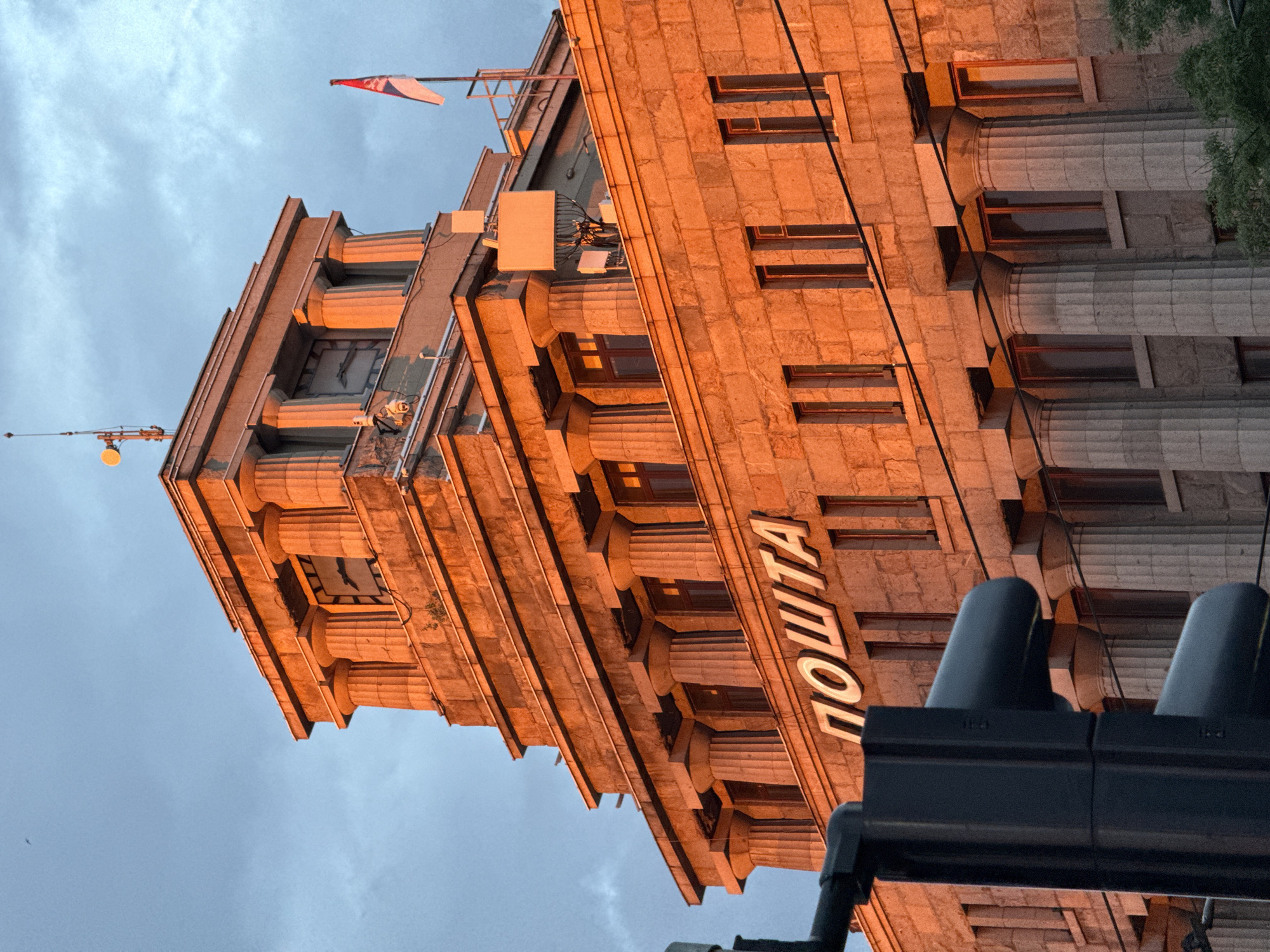 The image shows the facade of a stone building with classical architectural elements, likely the main post office in Vračar Municipality, Serbia. The building is illuminated by the warm, orange glow of the setting sun, creating strong highlights and shadows. Large columns line the facade, and there are visible windows. Prominently displayed on the building are the Cyrillic letters "ПОШТА", which translates to "Post Office". A Serbian flag is visible flying from a flagpole on the roof, as are various antennae and communication equipment. In the foreground, a traffic light is partially visible, indicating an urban setting. The sky is a mix of blue and grey clouds. There are no people or animals clearly visible in the image.