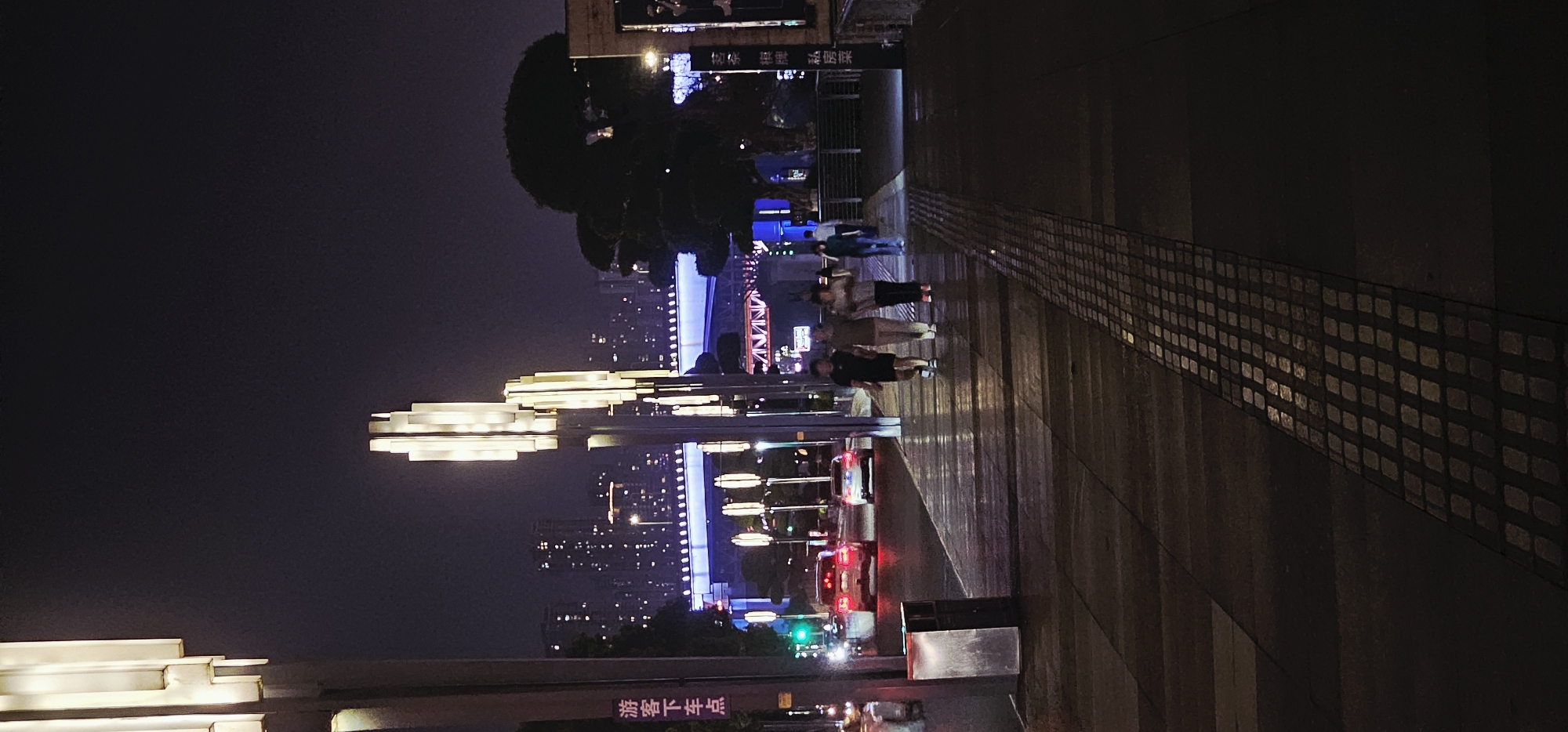 Captured at night in 江北城街道, China, the image depicts a street scene. Illuminated tiered light fixtures stand tall along the side of the road, contrasting against the night sky. Across the street, the city skyline emerges, speckled with lights, and a bridge is illuminated in blue. Several people stroll along the pathway, their forms slightly blurred due to the low light conditions. A tiled pathway lines the edge of the road, and traffic lights are visible, indicating the flow of vehicles. A trash receptacle is situated on the right side of the frame. A sign with Chinese characters is visible at the bottom of the image.