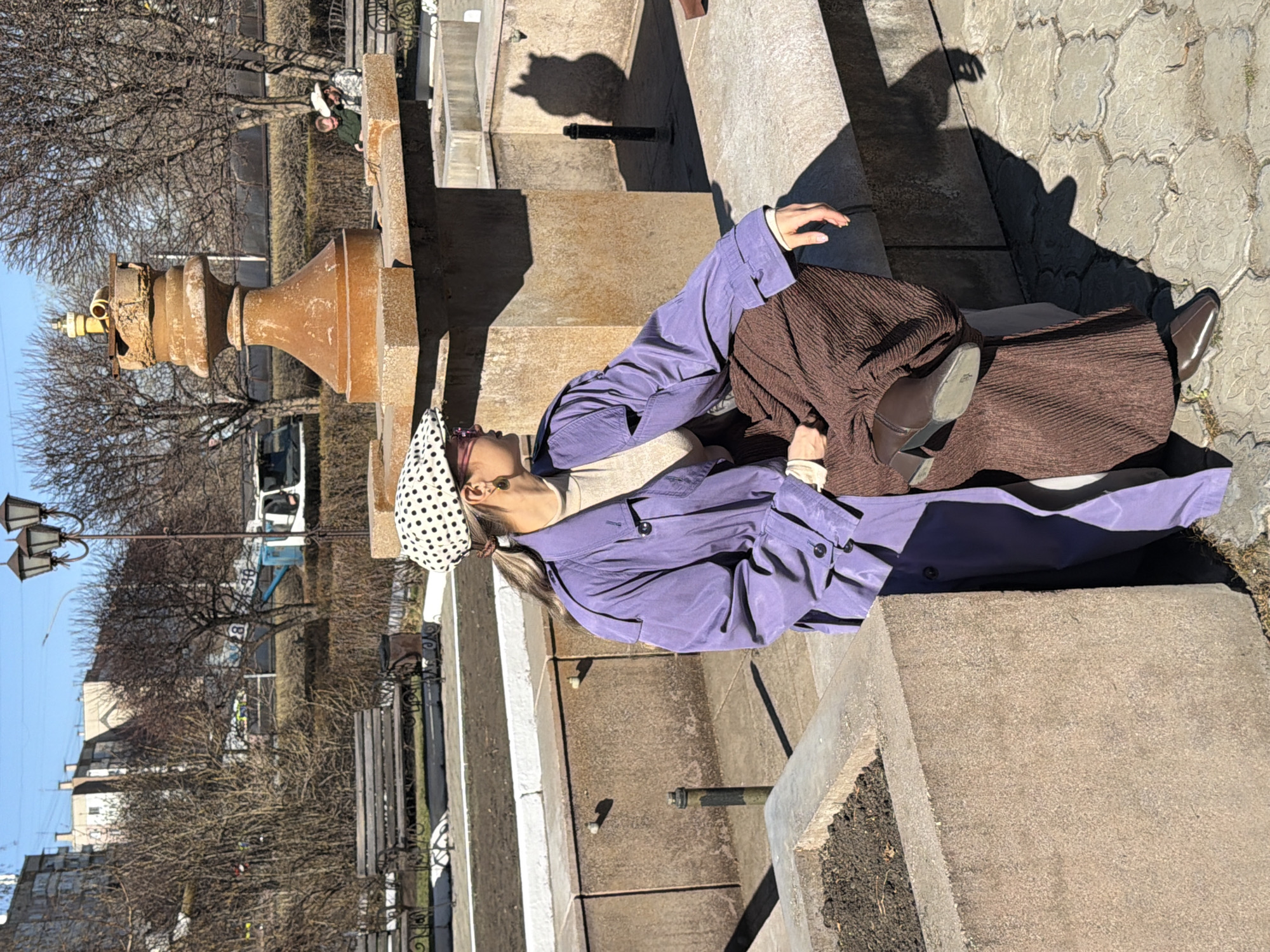 The original image is rotated 90 degrees clockwise, capturing a person resting or posing on concrete structures in an urban outdoor setting on a sunny day. The central subject, appearing female, wears a white and black polka-dot headscarf, a light-colored top, a long, light purple trench coat, and a long pleated brown skirt, complemented by brown shoes.
The setting appears to be a city park or public square in Krasnoyarsk, Russia. Various light brown concrete blocks and ledges form part of the landscaping, possibly an architectural feature or fountain base. To the person's left, a weathered, ornate, rusty metal object with a yellow top stands prominently. The bright sun casts distinct, long shadows of the person and objects onto the light gray, patterned pavement, indicating a clear sky.
In the background, bare trees suggest late autumn, winter, or early spring. A multi-headed black street lamp, wooden benches, and distant urban buildings with blurry vehicles, including a white van, are also visible. Small patches of dark soil are present in concrete planters. Two short metal pipes are embedded in one of the concrete surfaces. The person's pose is relaxed, and her gaze is directed away from the camera. While some faint numbers (e.g., "8", "39") are indistinctly visible on a pole in the background, no clear or fully legible text is present.
