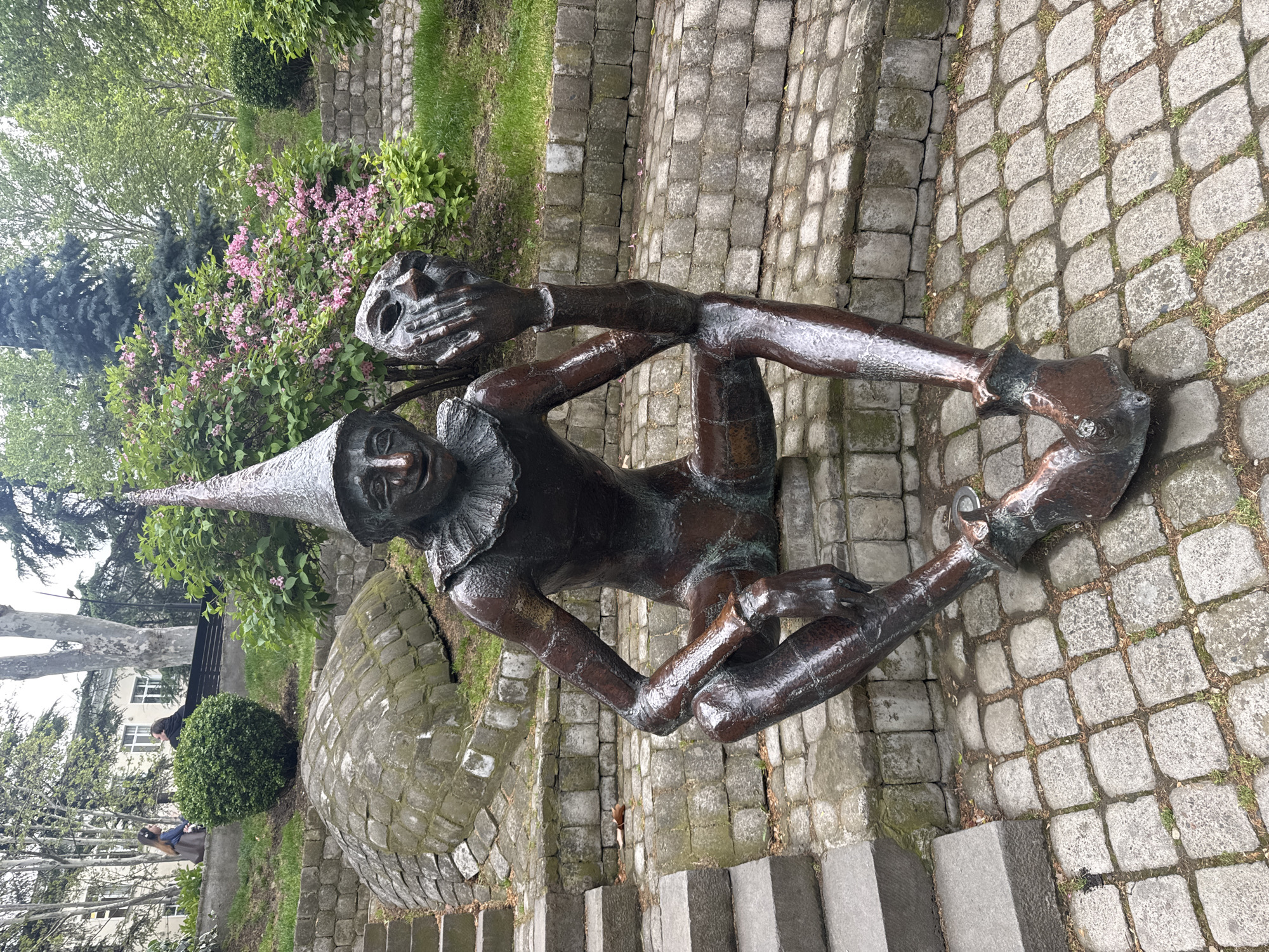 The image depicts a bronze sculpture of Rezo Gabriadze's "Master," a character in his puppet shows, located in Tbilisi, Georgia. The sculpture is positioned on a stone-paved area with steps leading to it. The figure wears a pointed hat and a ruffled collar and is seated in a relaxed pose, holding a theatrical mask in one hand. Behind the sculpture is greenery, including flowering bushes and trees. In the background, other buildings are visible. People sitting on a park bench can be seen in the back left.