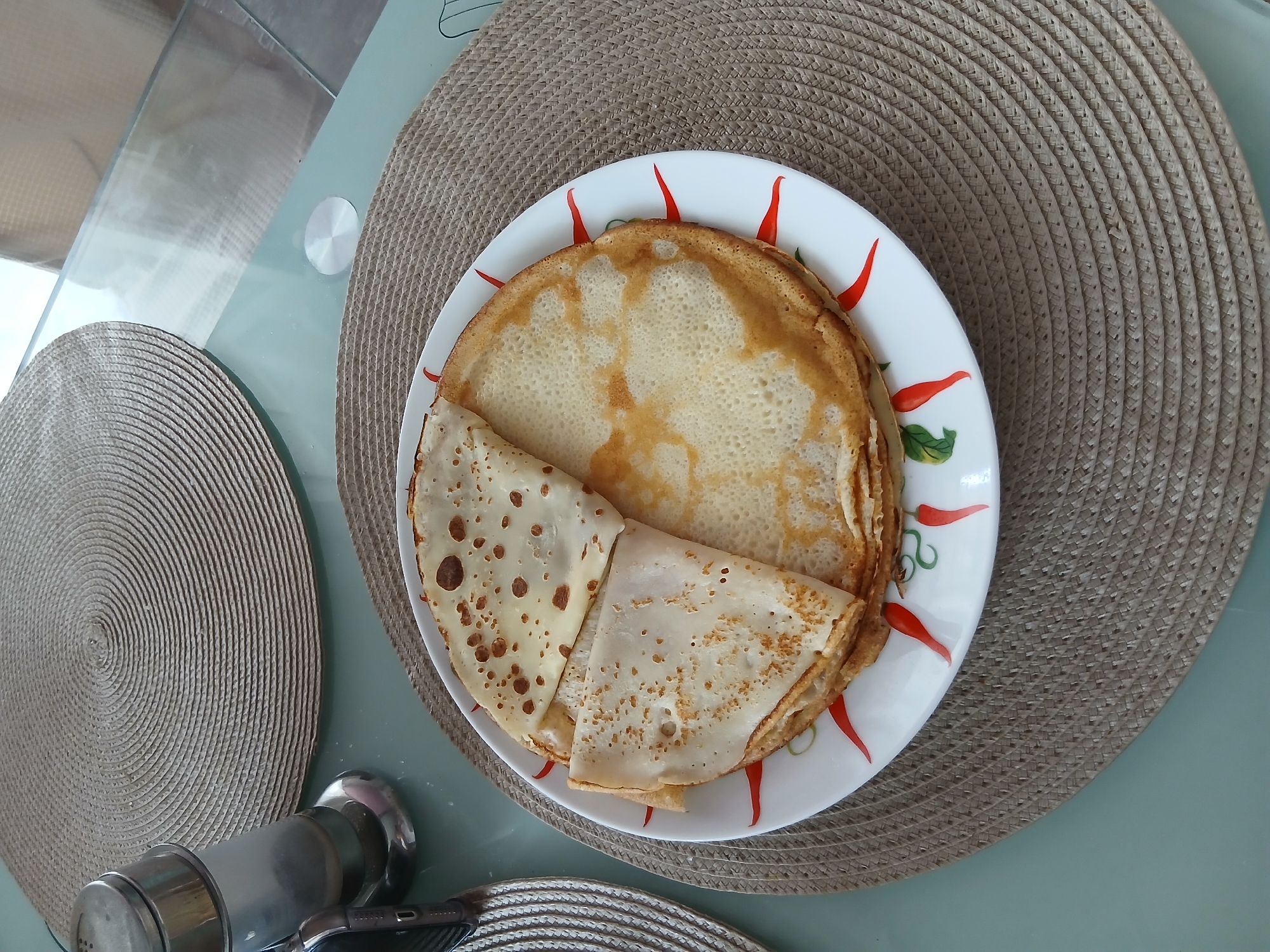 This media file is a top-down shot of a plate of crepes on a table. The crepes are stacked and folded on a white plate with red and green chili pepper decorations around the rim. The plate sits on a round, woven placemat with a textured, neutral color. To the left of the plate is another similar placemat. In the foreground, a metal salt and pepper grinder set is visible, along with a part of a smartphone. The table surface appears to be glass, reflecting the light. The scene suggests a casual meal or snack. There is no visible text in the image. The lighting suggests it is daytime. The presence of a salt and pepper grinder and a phone implies this is a domestic setting, likely a kitchen or dining area. The location context of Chaikovsky, Russia, further solidifies this as a personal, everyday scene.