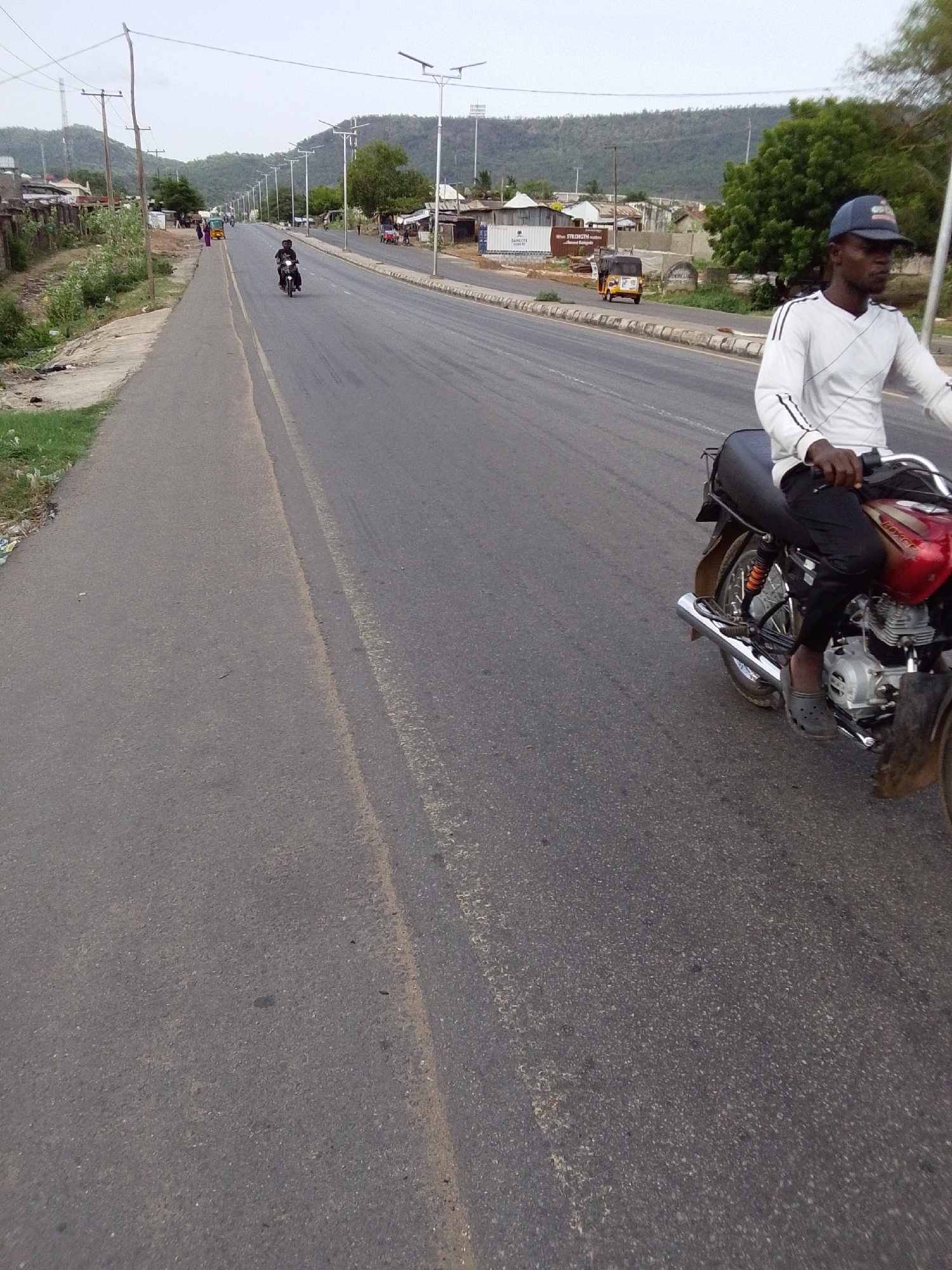The image depicts a man riding a red motorcycle on a paved road in Adankolo, Nigeria. He wears a long-sleeved white shirt, dark pants, and a blue baseball cap. Another motorcyclist is visible further down the road. Buildings and vegetation line the roadside. In the background, distant hills and utility poles are present. The sky appears overcast. A small, yellow three-wheeled vehicle is also visible down the road.