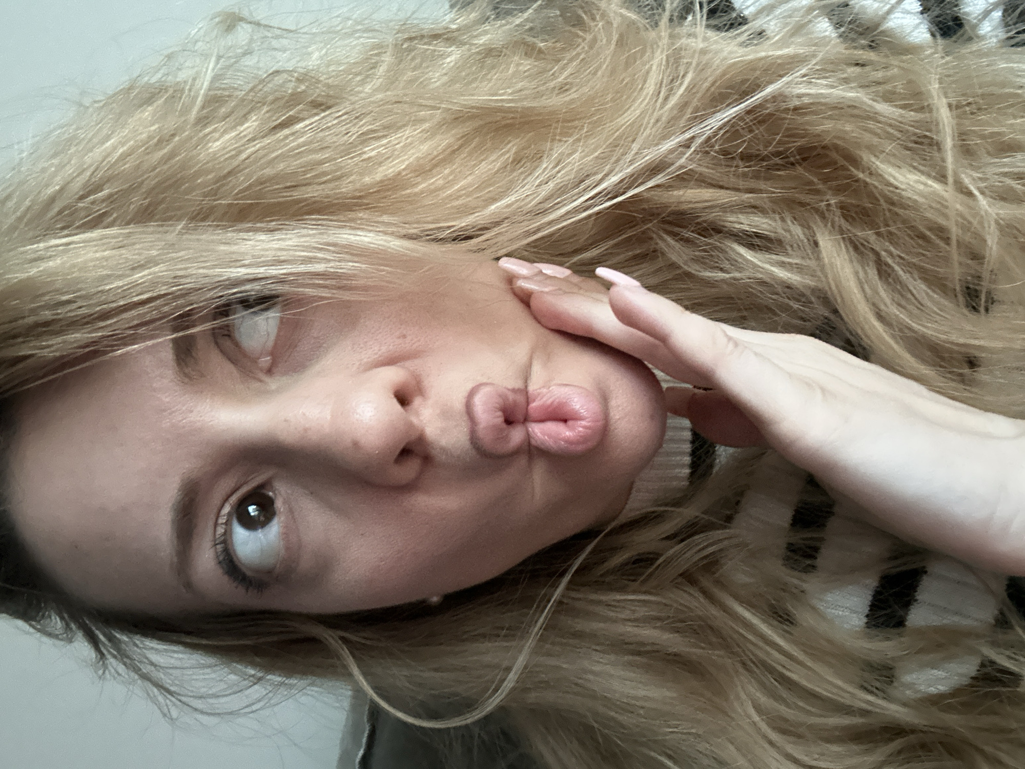 A close-up, tilted indoor shot features a woman making a playful "fish lips" or puckered face. Her head is angled to the left, with her dark brown eyes looking upwards, enhancing the whimsical expression. She has long, flowing, wavy blonde hair that dominates much of the frame, showing various shades of blonde and light brown. Part of her left hand is visible on the right side of the image, gently touching her face and hair, revealing light pink manicured fingernails.
She is wearing a garment with a black and white striped pattern, partially visible beneath her hair and arm. The background is a simple, light-colored surface, indicating an indoor setting. The lighting is bright and appears even, suggesting good indoor illumination. The image conveys a playful and lighthearted emotion. No text is visible in the media file. The provided location context for this scene is Amsterdam, Netherlands.