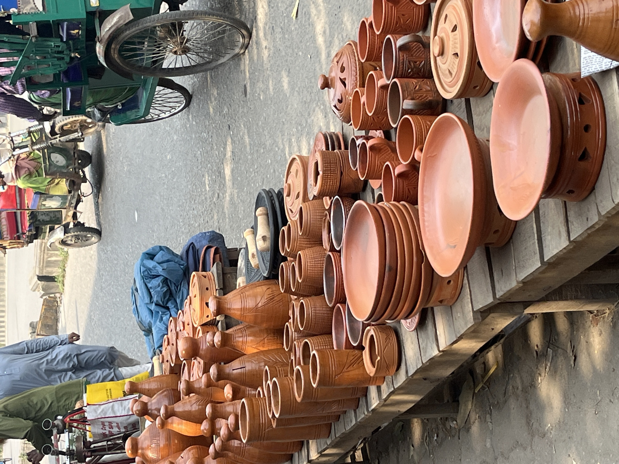 The image depicts a street scene in Dhaka, Bangladesh, with a focus on a display of earthenware pottery for sale. A variety of pottery items, including vases, bowls, cups, and lidded containers, are arranged on a makeshift wooden shelf. The pottery is predominantly a reddish-brown color with decorative patterns. In the background, there are vehicles and people on the street. A blue tarpaulin covers some of the pottery. To the left, a man in a blue shirt is visible, as well as a man in a green shirt. The environment appears to be an outdoor market or roadside vendor setup.