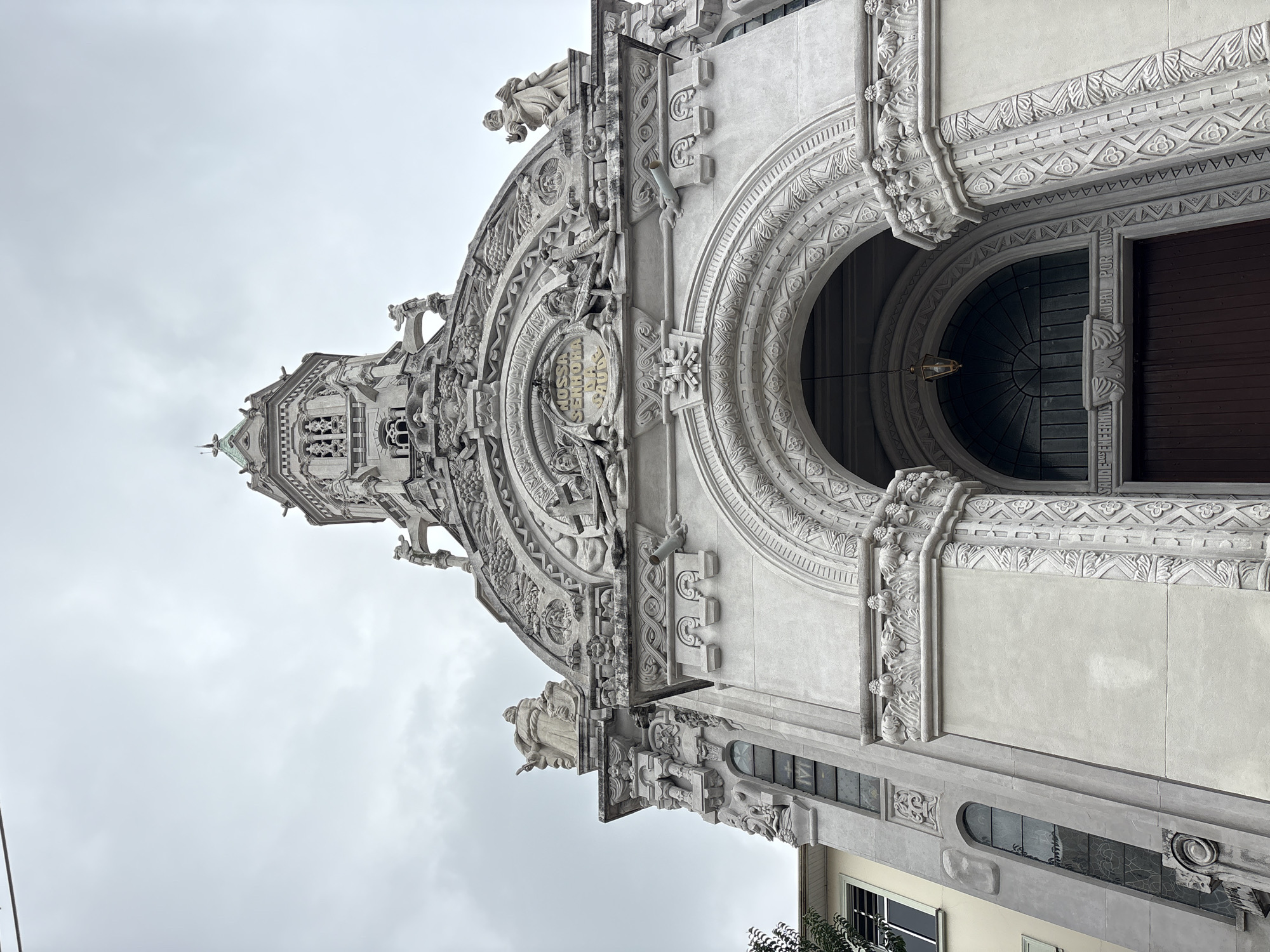 The image shows a low-angle, close-up exterior view of the front facade of the Santuário Nossa Senhora de Fátima, a Catholic church located in the Pompeia neighborhood of São Paulo, Brazil. The sky is overcast and appears gray. The church's architecture is ornate, featuring intricate stone carvings, arched doorways, and statues. At the top of the facade, a detailed tower with a green spire rises into the sky, adorned with decorative elements and smaller statues. Below the tower, a large arched entrance is visible, with a dark wooden door and a lantern hanging above it. The building's facade is made of light-colored stone and exhibits a significant amount of sculpted detail, including relief carvings of figures, foliage, and decorative patterns. The left side of the building shows a section of an adjacent building with a yellowish facade and a few windows. The overall impression is of an elaborate and aged religious structure.