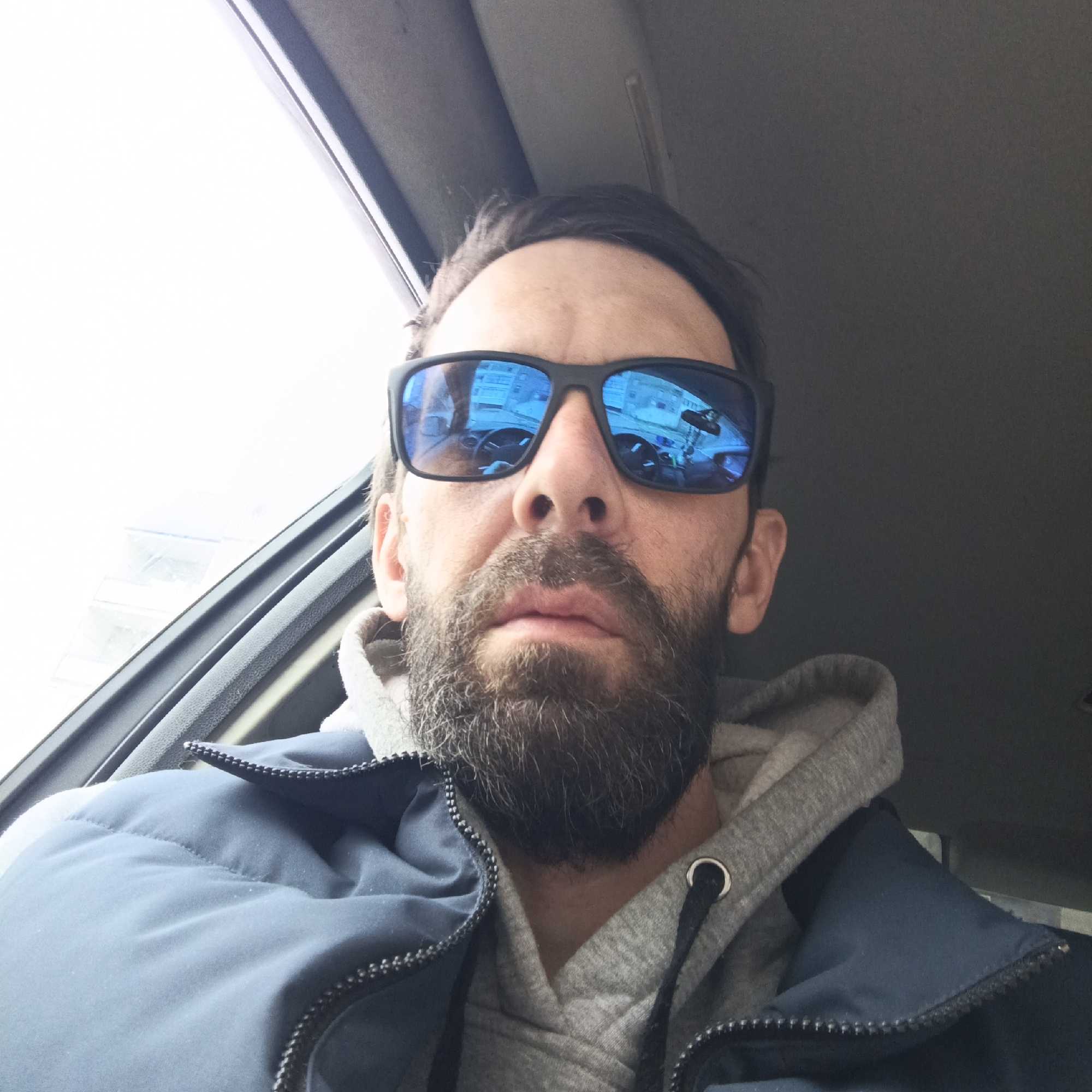 A man with a beard and wearing sunglasses and a hooded sweatshirt is sitting in the driver's seat of a car. The reflection in the sunglasses suggests the man is in Марковский, Russia.