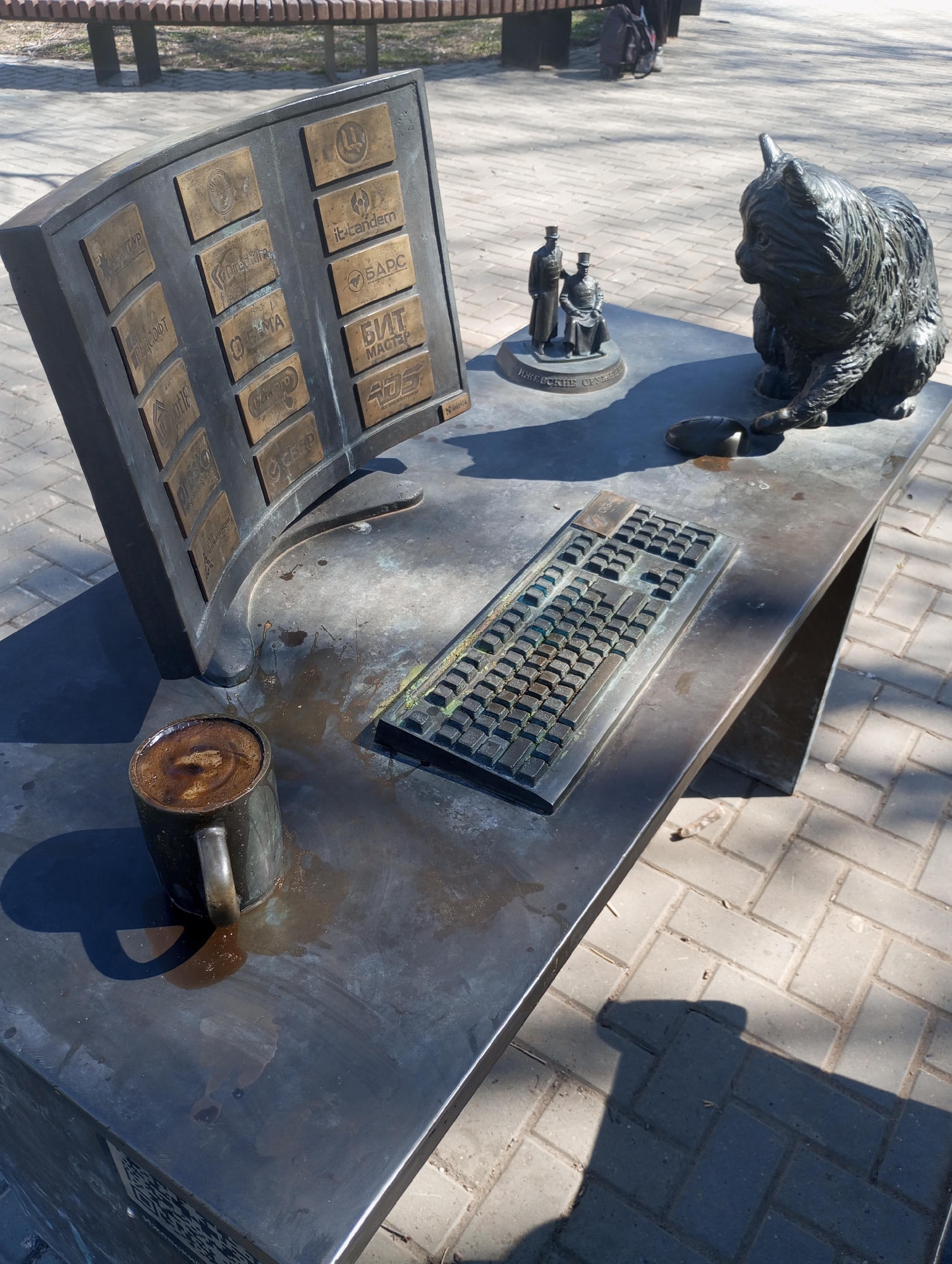 The image shows a metal sculpture in Pervomaysky, Russia. The sculpture depicts a desk with a curved computer monitor displaying several company logos. There is a keyboard on the desk, a bronze sculpture of two men, a cat statue, a spilled coffee cup, and coffee stains. The entire setting appears to be an outdoor monument on a paved area.