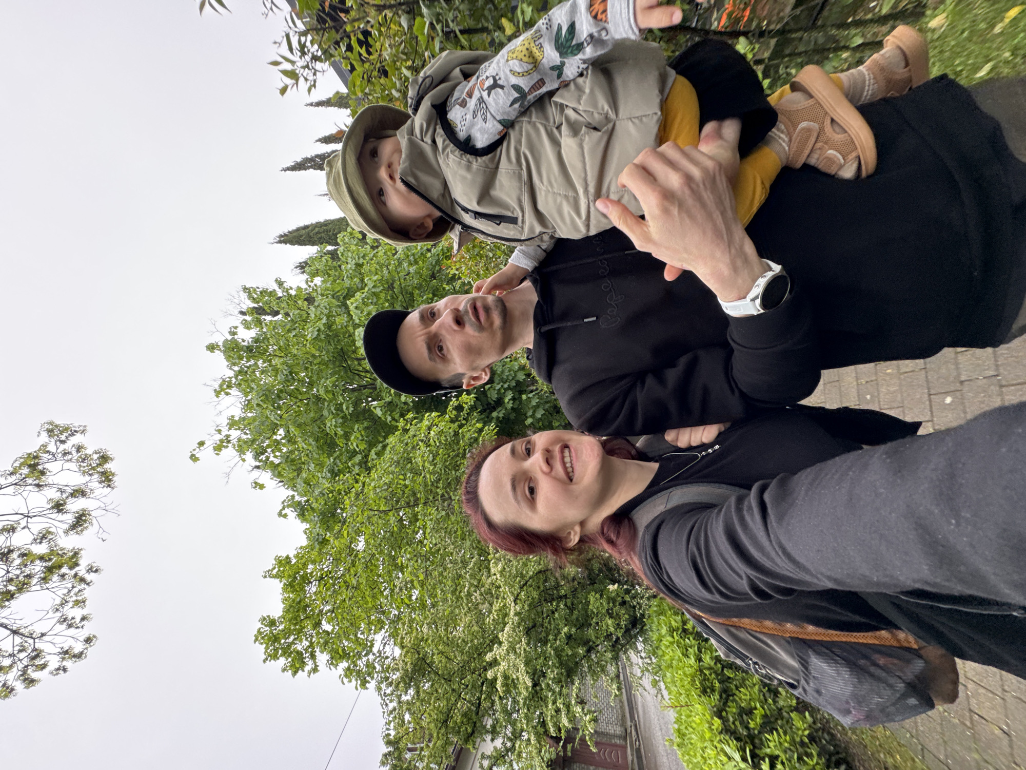 A family stands on a paved path, potentially in Sochi, Russia, amidst lush greenery and trees under an overcast sky. The image is taken from a low angle, capturing a woman smiling at the camera, holding a phone or camera to take a selfie. Next to her is a man wearing a black cap and black sweatshirt, holding a small child with fair skin and blonde hair, dressed in a light-colored jacket, yellow pants, and a matching hat. The child is sitting on the man's shoulders. A building and a few trees are visible in the background.