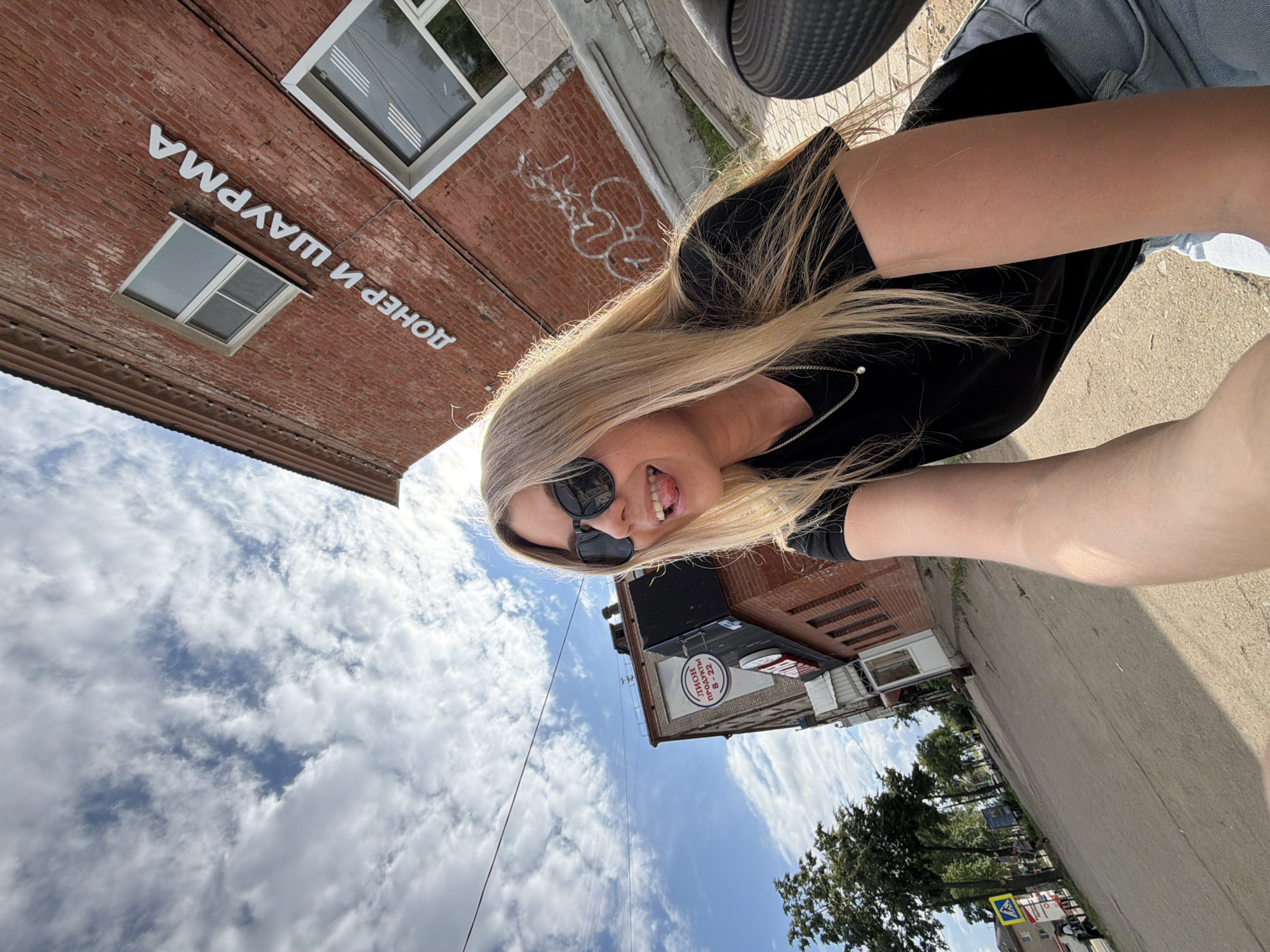 The image captures a selfie taken from a low angle by a young blonde woman with sunglasses, sticking her tongue out. She is wearing a black t-shirt and blue jeans. Behind her, a brick building with white lettering that reads "ДОНЕР И ШАУРМА" (Doner and Shawarma) is visible, along with windows. To the left, another building has a circular sign with "ЛИОВ ПРОДУКТЫ 8-22" (Liov Products 8-22). The scene appears to be on a street in Chaikovsky, Russia, with a sidewalk, trees, and a traffic sign. The sky is cloudy, with patches of blue.