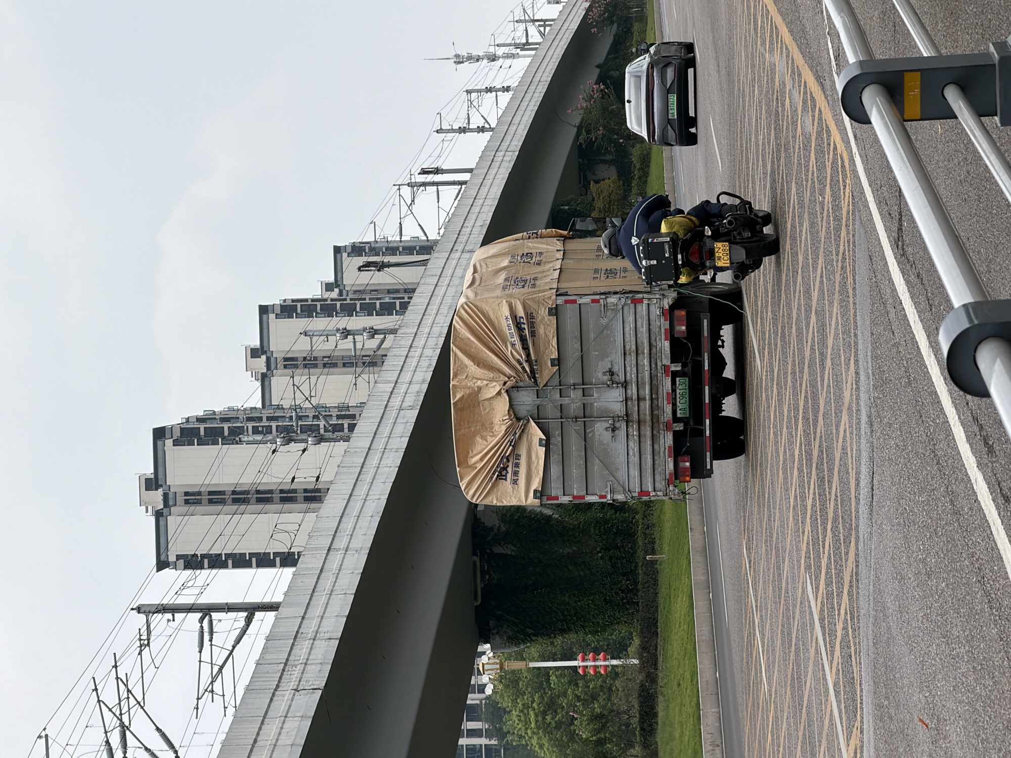 This image depicts a street scene in Jiangjin District, Chongqing, China, featuring a multi-lane road with a truck and a motorcycle. The truck, covered with a beige tarp, is in the foreground, with its rear license plate visible as "A C96130." A person on a motorcycle, also with a visible license plate "1C088," is riding in the lane to the right of the truck. A dark-colored sedan is visible further down the road on the right. Overhead, a large elevated structure, likely a highway or railway, supported by concrete pillars, dominates the background. The pillars are covered in dense green ivy. Buildings with multiple stories are visible behind the elevated structure, and overhead power lines stretch across the sky. The road surface is asphalt, marked with yellow grid lines indicating a no-parking zone. The sky is overcast.