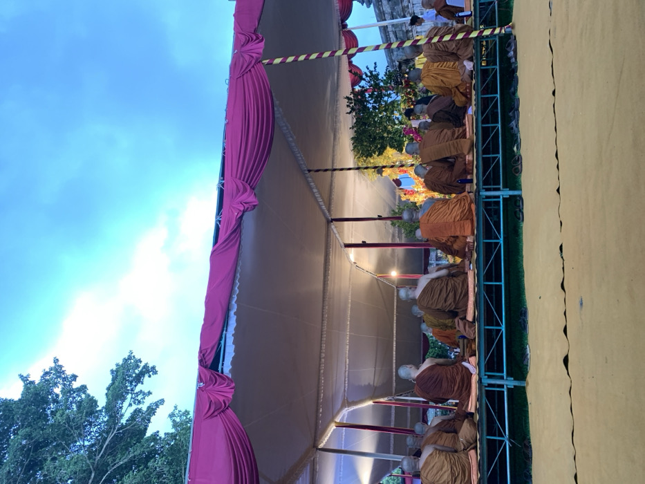 A group of 15 Buddhist monks are seated facing a large, elaborately decorated altar in a large tent structure. The tent has a white fabric roof and pink fabric drapes lining the front and sides. It is located in the city of Mungkid, Indonesia at coordinates -7.605, 110.23. There are a number of people seated on benches in front of the monks, with one person in a bright green shirt and orange pants and another standing, wearing a brown robe. The monks are wearing brown robes and are sitting with their legs crossed. There are a few green plants visible on the ground, as well as a large green tree in the background. The sky is blue with white clouds.
