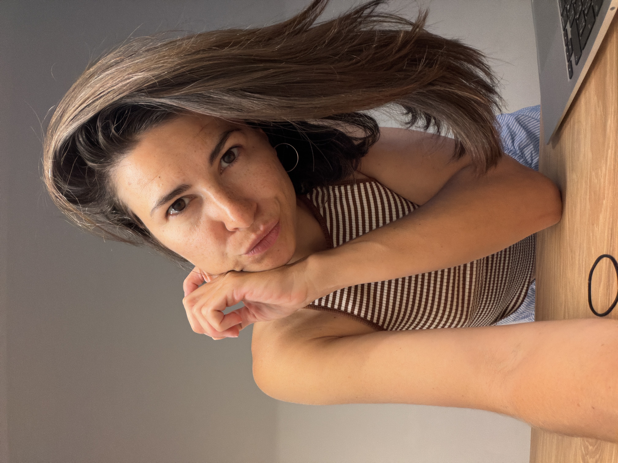 A woman with long brown hair, fair skin, and brown eyes is positioned in the foreground, tilting her head towards the camera. She is wearing a striped top, with one arm resting on a wooden surface, likely a desk. A laptop is visible in the top right corner of the frame. A black hair tie is on the wooden surface. The background consists of a plain grey wall. No other details suggest a specific location in Budva, Montenegro.