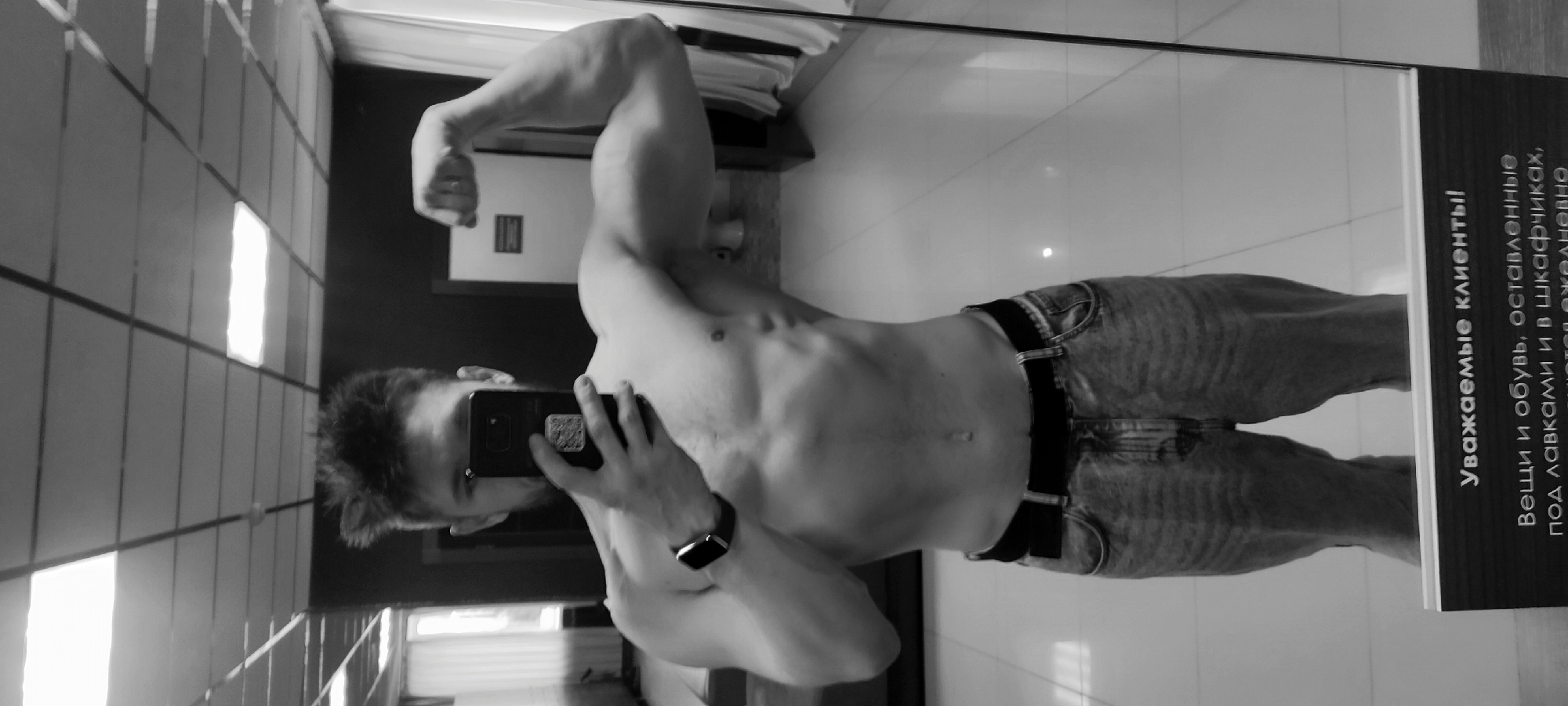 A black and white medium shot captures a shirtless man flexing his bicep in what appears to be a mirror selfie. He is wearing jeans with a black belt and an Apple Watch on his left wrist. The background includes a tiled wall, a closed white door with a sign, and another sign with text in Russian that reads "Уважаемые клиенты! Вещи и обувь, оставленные под лавками и в шкафчиках, пожалуйста ждановно," which translates to "Dear customers! Please leave your belongings and shoes under the benches and in the lockers." Based on the text and general aesthetic, the location is likely a gym or a similar facility in Russia. The image is oriented sideways, requiring rotation for proper viewing.