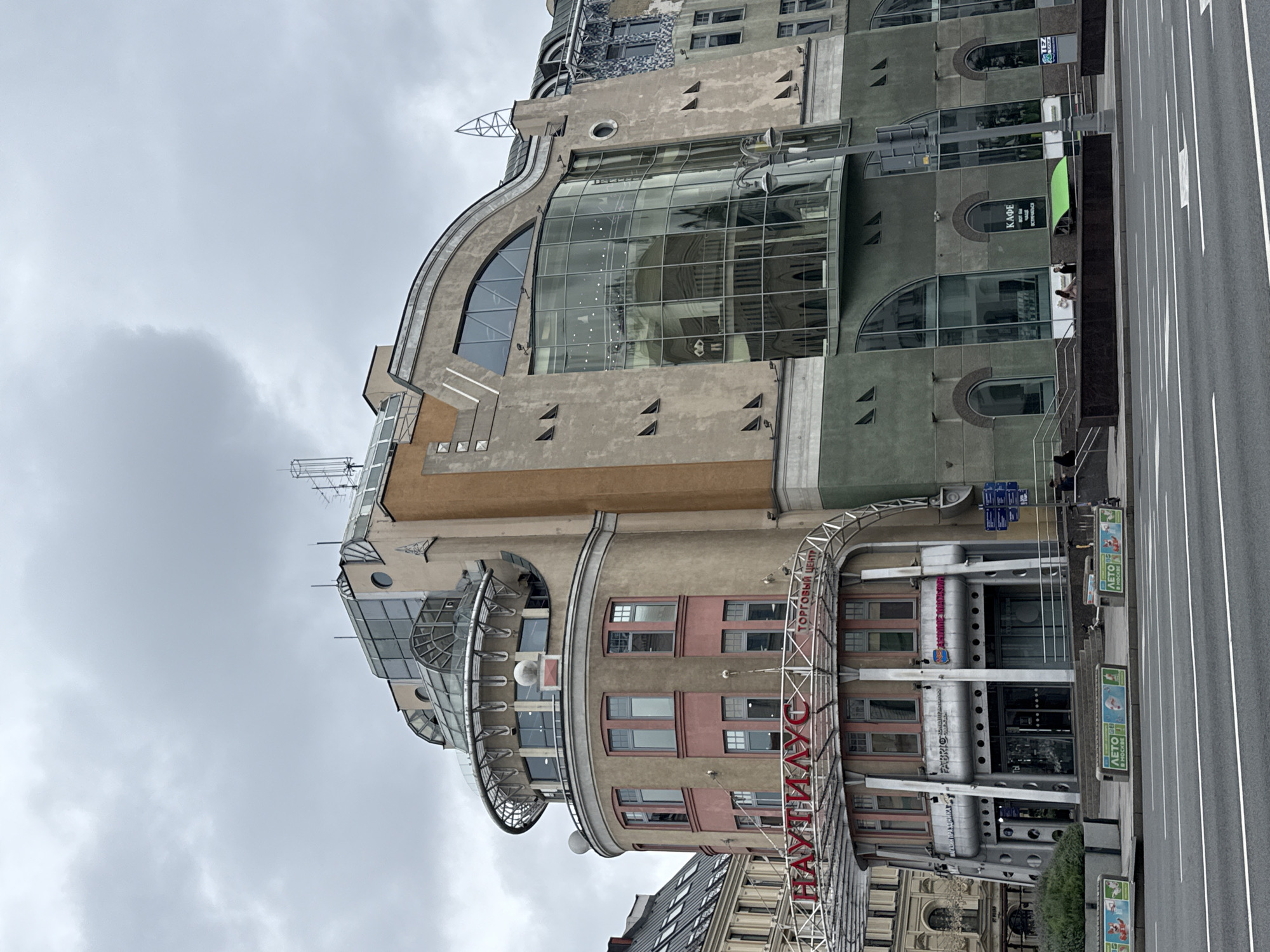 The image shows a building in Moscow, Russia, identified by the sign "НАУТИЛУС" which translates to "Nautilus" and "ТОРГОВЫЙ ЦЕНТР" which means "Shopping Center". The building has a distinctive architectural style, featuring a circular tower with a pink and beige facade, topped by a glass dome and a metal railing structure. Adjacent to the circular part is a larger section with a curved, multi-story glass facade, reflecting the interior and the sky. The building's exterior also includes sections with a taupe color, a moss green lower section, and some decorative elements. Multiple signs are visible on the building, including store names and directions. In the foreground, a street with white lane markings is present, with some concrete planters and advertising banners displayed along the sidewalk. A few people are visible walking on the sidewalk and near the building entrance. The sky is overcast and gray.