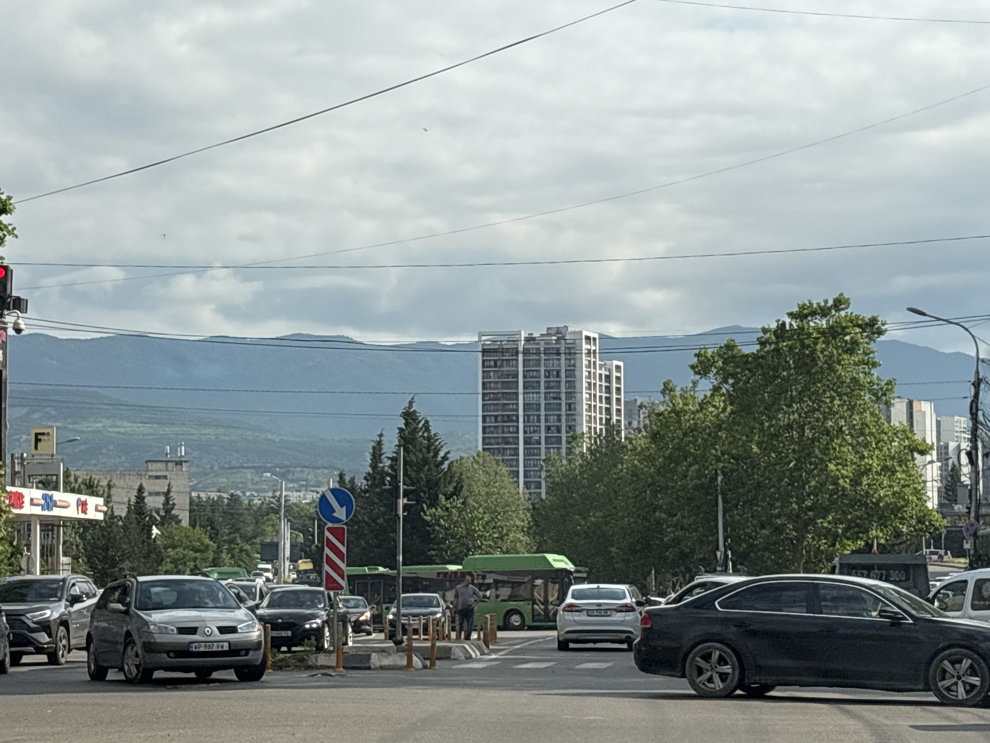 The image depicts a traffic scene in Tbilisi, Georgia, likely on a cloudy day. In the foreground, several cars are stopped or moving slowly at an intersection. A silver hatchback car with license plate "WP 987 PW" is prominent in the center, with other vehicles including a dark sedan and an SUV visible. A pedestrian stands near a crosswalk, next to a traffic light and a blue directional sign. In the middle ground, green buses are stopped, with another pedestrian standing near one of them. Behind the buses, a tall, modern white building with many windows stands out against the sky. The background features lush green trees and distant blue mountains, indicative of a city with surrounding natural landscapes. Power lines are visible crisscrossing the sky. A traffic light with a red light illuminated is visible on the left side of the image.