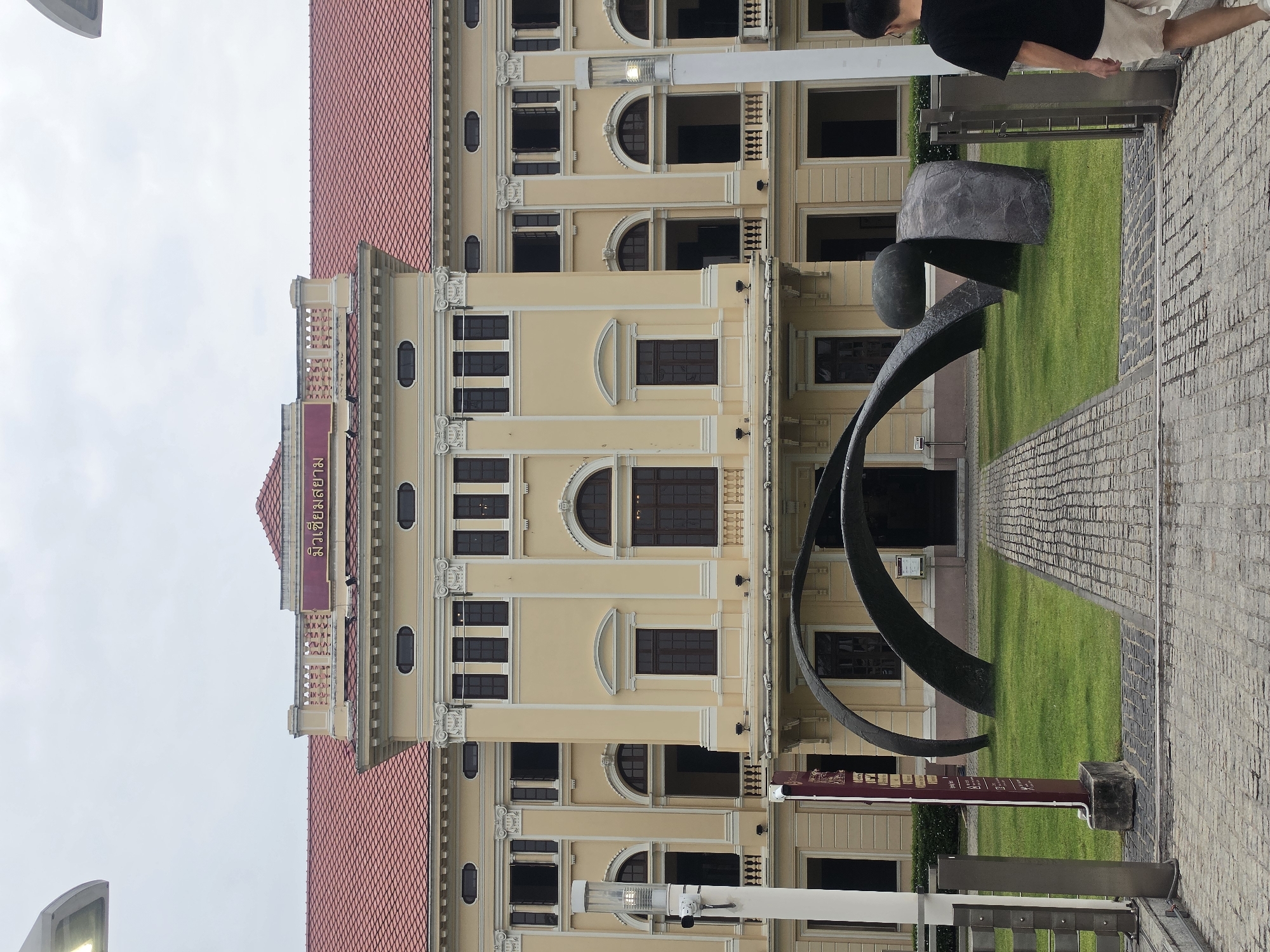 The image shows the Faculty of Fine and Applied Arts building at Thammasat University in Bangkok, Thailand. The building is a multi-story structure with a light yellow facade, a red-tiled roof, and multiple windows and architectural details. In front of the building is a green lawn with a modern art sculpture made of dark metal. A pedestrian walkway made of light-colored brick extends from the sculpture towards the viewer. A person is partially visible at the top right of the image, standing on the walkway. A signpost with information about the location stands near the sculpture. The sky is overcast.