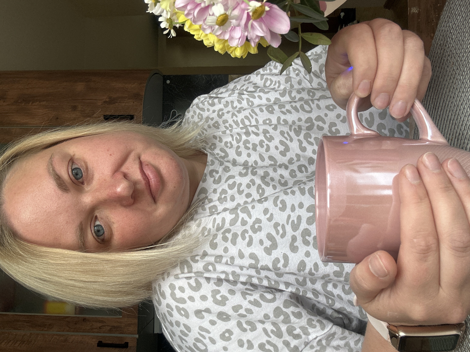 A slightly smiling blonde woman with light blue eyes looks directly at the camera in this indoor portrait, which is rotated 90 degrees counter-clockwise. She has fair skin and shoulder-length hair. The woman is casually dressed in a grey and white leopard print top. She holds a light pink ceramic mug in both hands, and a smartwatch with a light-colored band and metallic casing is visible on her left wrist. Her fingernails appear natural and unpolished.
Behind her, a bouquet of flowers, including pink, yellow, and white blossoms with green foliage, can be seen in the upper right. The background on the left features wooden furniture, possibly a dresser or cabinet, with dark handles. A dark, possibly electronic, object is also visible behind her head. The lighting is soft, suggesting a casual, relaxed moment, possibly at home. No text is visible in the image. The setting is in Chaikovsky, Russia.