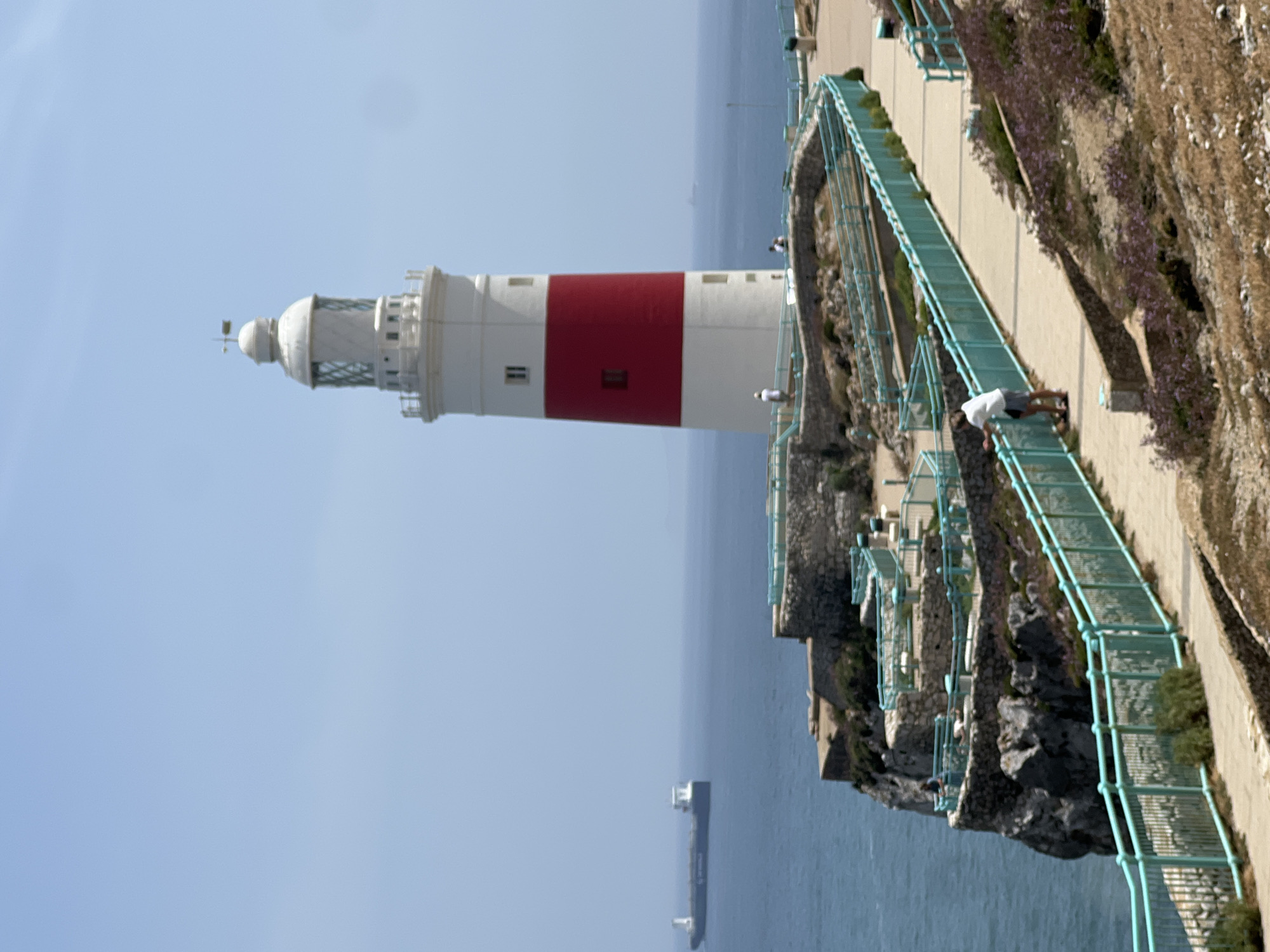 This is a photograph of the Europa Point lighthouse, located at the southernmost tip of Gibraltar. The lighthouse, a prominent white and red striped tower, stands tall against a clear blue sky. In the foreground, a paved pathway winds along a cliff edge, bordered by a light teal railing. Several people are visible on the pathway and on tiered stone platforms surrounding the lighthouse. Some are walking, while others are standing and looking out at the sea.
The sea is a deep blue and stretches to the horizon. A large cargo ship is visible in the distance on the left side of the frame, its silhouette dark against the water. The cliff face is rugged and rocky, with sparse vegetation. The overall scene conveys a sense of openness and a panoramic view of the coastline and the sea. The lighting suggests it is a sunny day.