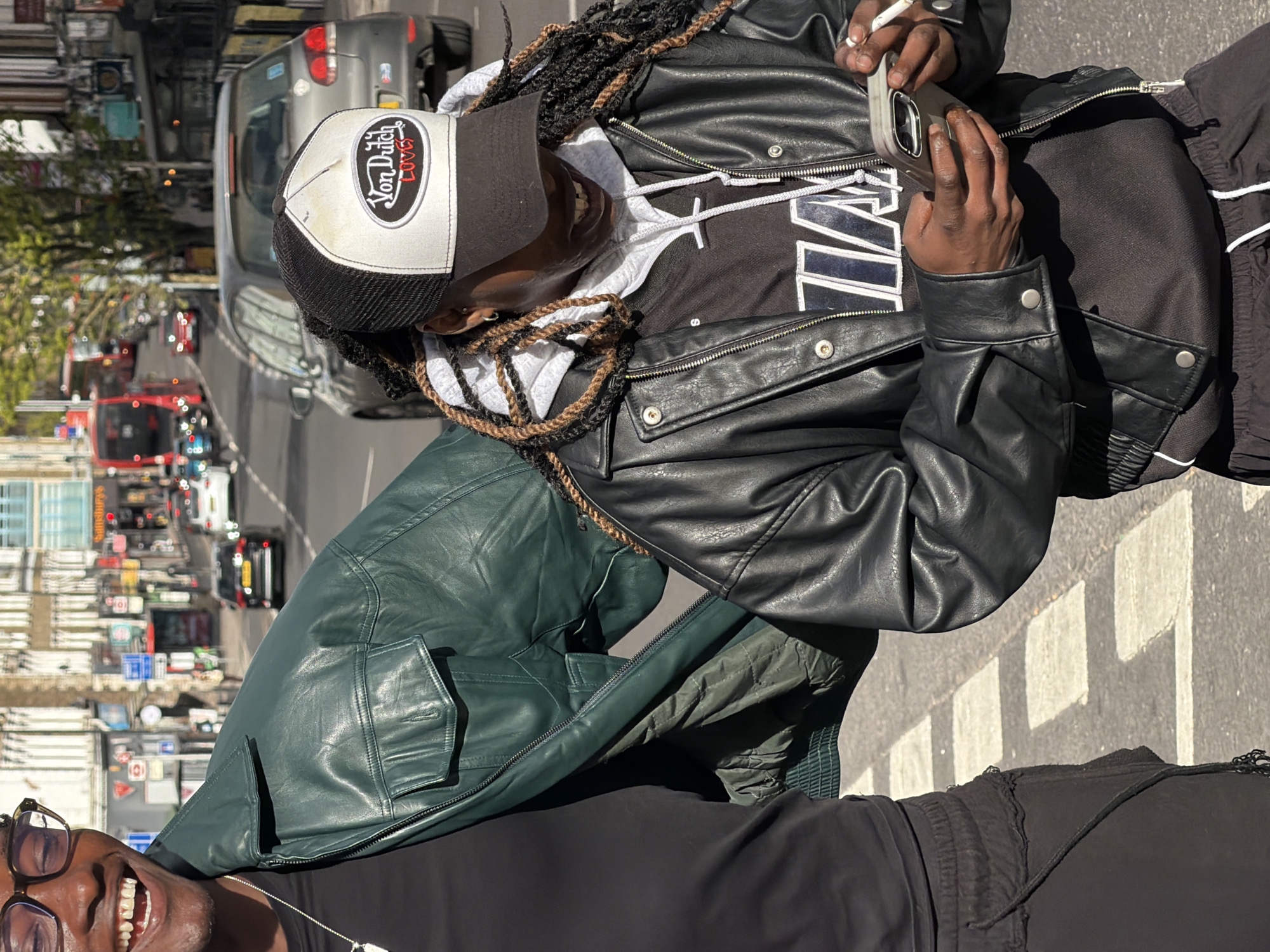 Two individuals are visible in an urban setting, likely London, United Kingdom, given the visible street signage and red buses. The primary subject, situated at the top of the frame, is holding a cigarette and a mobile phone, wearing a black and white cap, a black and grey hoodie, a black leather jacket, and black pants. Their hair is styled in braids with brown accents. A secondary individual, only partially visible at the bottom of the frame, is smiling and wearing glasses. They are dressed in black. A green leather jacket rests next to them. The background showcases a busy street with traffic, including red double-decker buses and cars, along with pedestrian crossings. Street signs and buildings provide context to the city location.