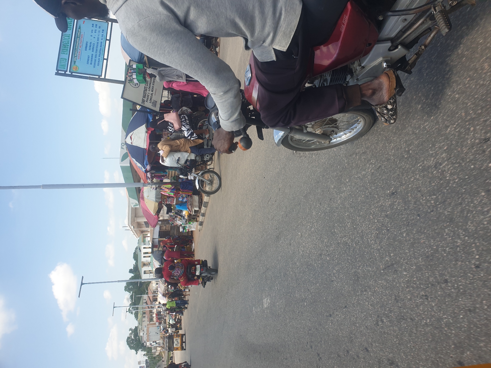 The image shows a view from a moving motorcycle on a street in Lokoja, Nigeria. The street is lined with shops and vendors, some shaded by umbrellas. Other motorcycles and pedestrians are visible on the road. Signage for the "United Evangelical Church" is present, located on UEC Road, Lokoja, along with another sign for the "Chief Magistrate Court Phase 1-Lokoja, Kogi State". The camera is positioned at an angle to the ground, showing the motorcycle’s handlebars and the rider's foot in a sandal resting on the footrest. The sky is partially visible.