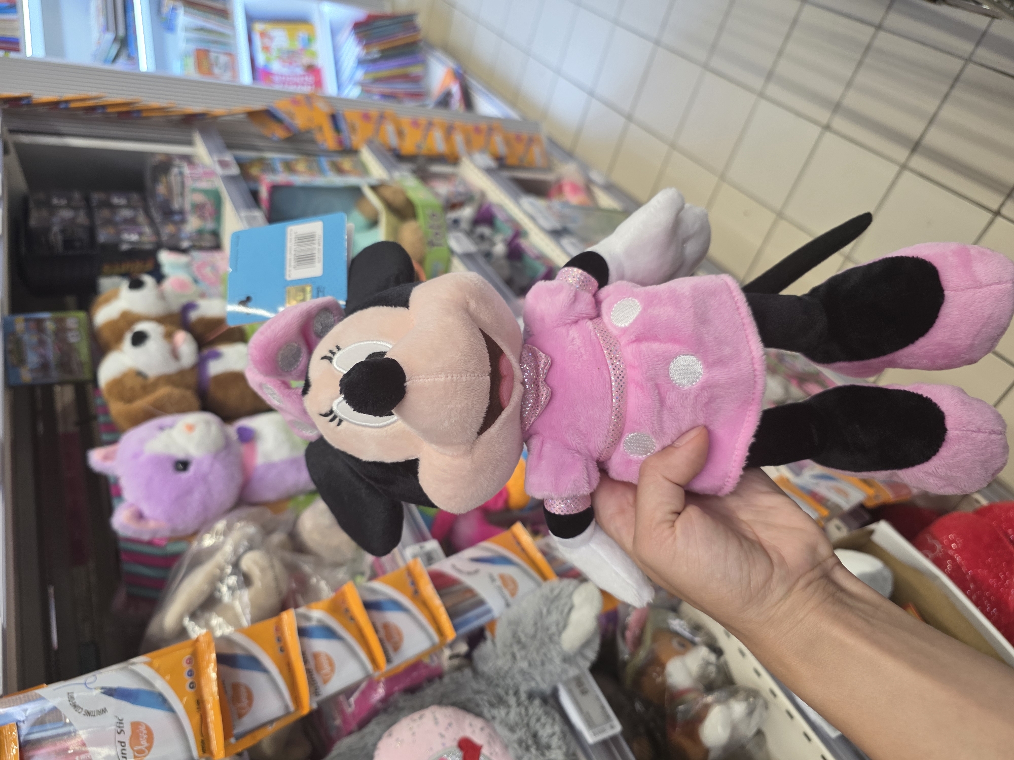 A hand holds a Minnie Mouse plush toy in a store. The toy is pink with white polka dots and a pink bow. In the background, shelves are stocked with various items, including other stuffed animals, pens, and books. The floor is tiled.