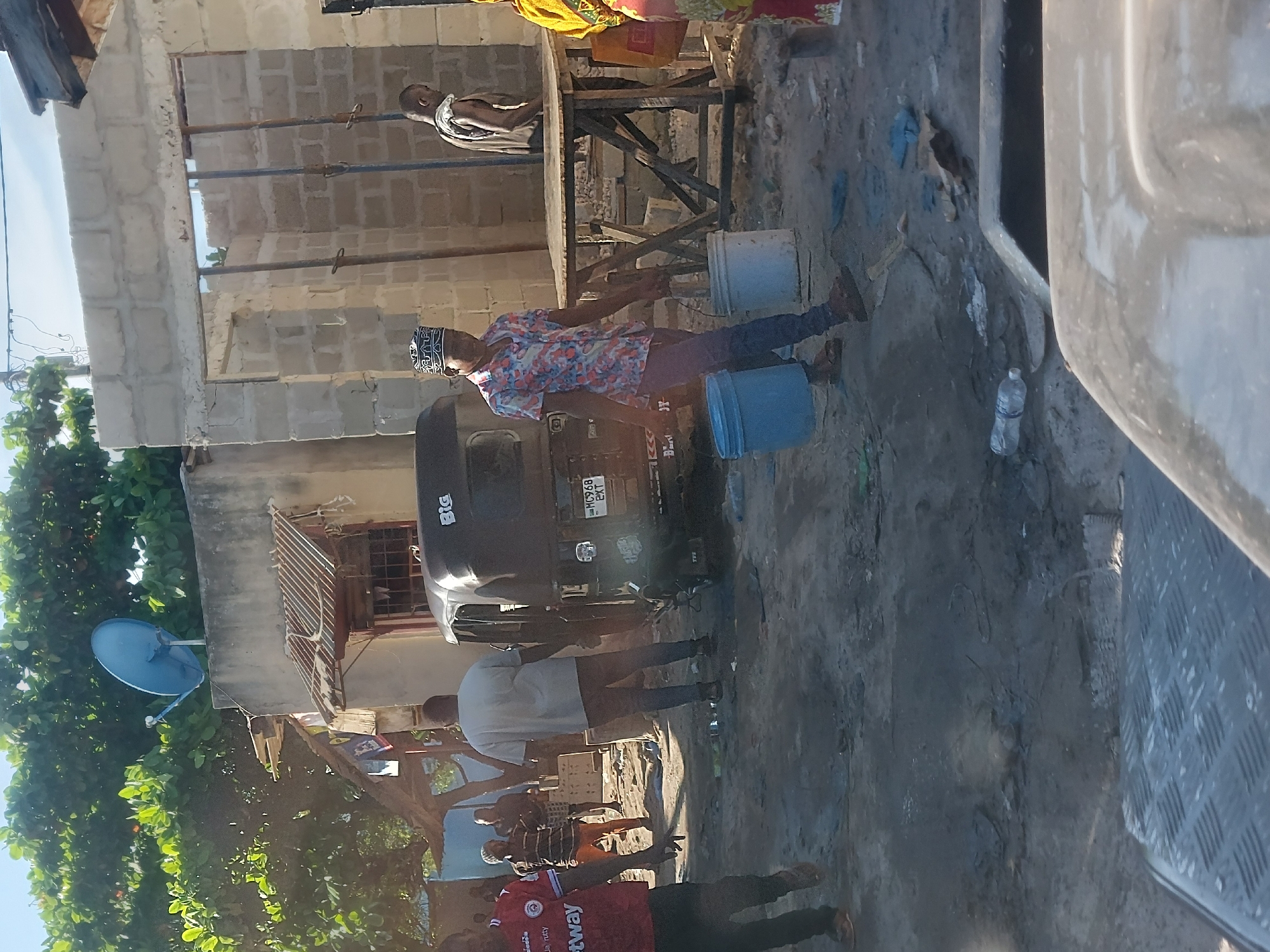 In Dar es-Salaam, Tanzania, a bustling street scene unfolds. A man wearing a patterned shirt and traditional cap carries a blue bucket, possibly filled with water, and interacts with a tuk-tuk style vehicle. Several other people are nearby, including one wearing a red shirt with a "Betway" logo. The scene is set against a backdrop of partially constructed brick buildings, wooden structures, and lush green trees. A blue satellite dish is affixed to one of the buildings. The foreground reveals a concrete surface with a discarded water bottle. Part of a vehicle is visible in the right side of the image.
