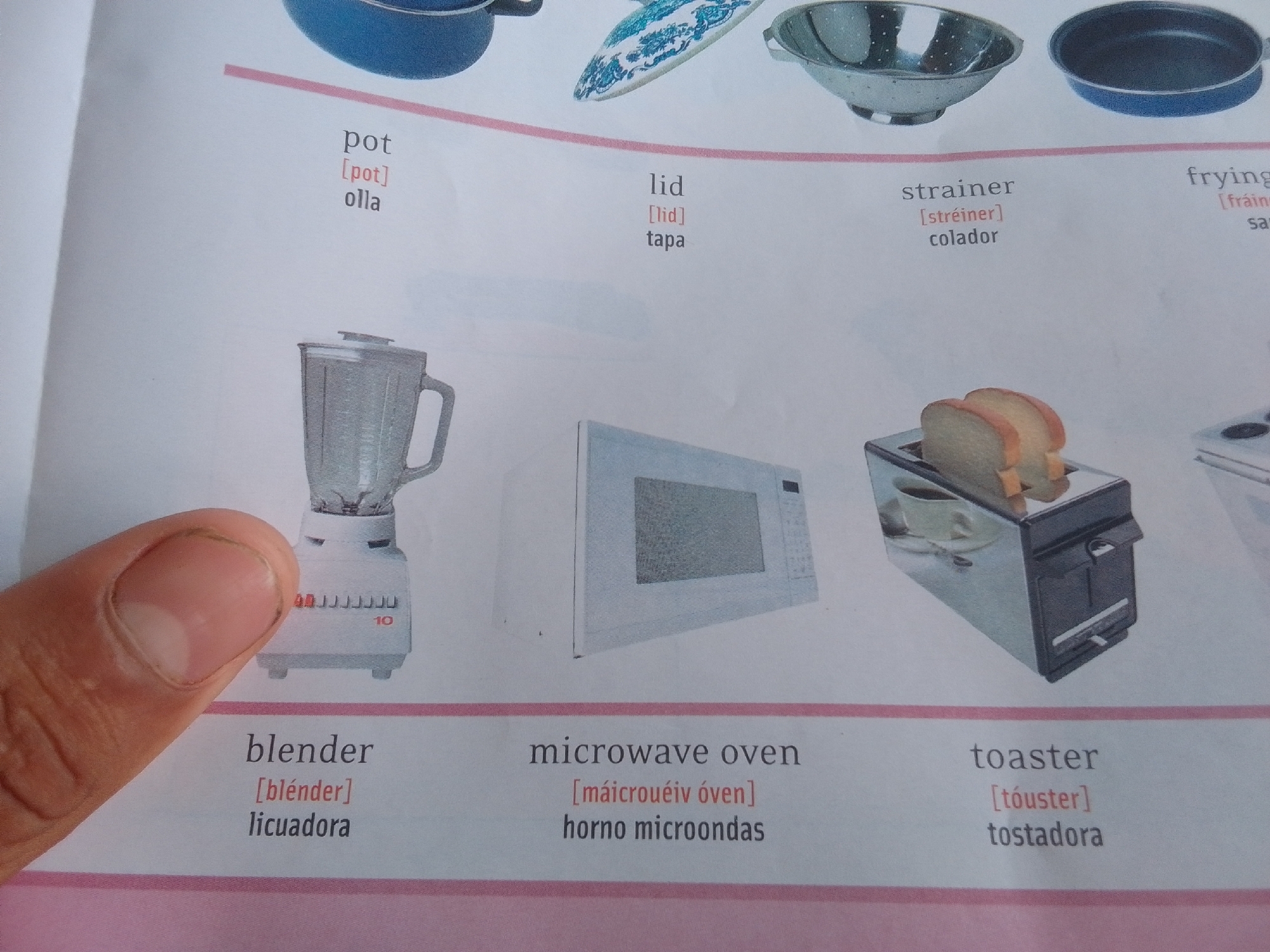 This media file is a close-up photograph of a page from a children's book or educational material, likely a vocabulary lesson about kitchenware. The page features illustrations and text in both English and Spanish.
In the upper portion of the image, several kitchen items are depicted: a blue pot, a patterned lid or dish, a metal strainer with holes, and a dark frying pan. Below these images, corresponding labels are provided in English, phonetic English, and Spanish. The words visible are "pot," "lid," "strainer," and "frying."
The lower half of the page displays three distinct kitchen appliances. On the left is a white blender with a clear jug and a dial that reads "10". Next to it is a white microwave oven. To the right, a chrome toaster holds two slices of bread, with a cup and saucer visible inside its opening. Below these appliances are their English and Spanish labels: "blender," "microwave oven," and "toaster."
A person's thumb and forefinger are visible in the bottom left corner of the image, obscuring part of the blender. The background is a plain white page. The overall setting is a brightly lit indoor environment, consistent with a book's page. There are no apparent weather cues or time of day indicators. The dominant text includes the words "pot," "lid," "strainer," "frying," "blender," "microwave oven," and "toaster," along with their phonetic pronunciations and Spanish translations. The location context provided is La Tagua, Venezuela, suggesting the origin or intended audience of the material. There are no emotional cues conveyed as the image focuses on inanimate objects.