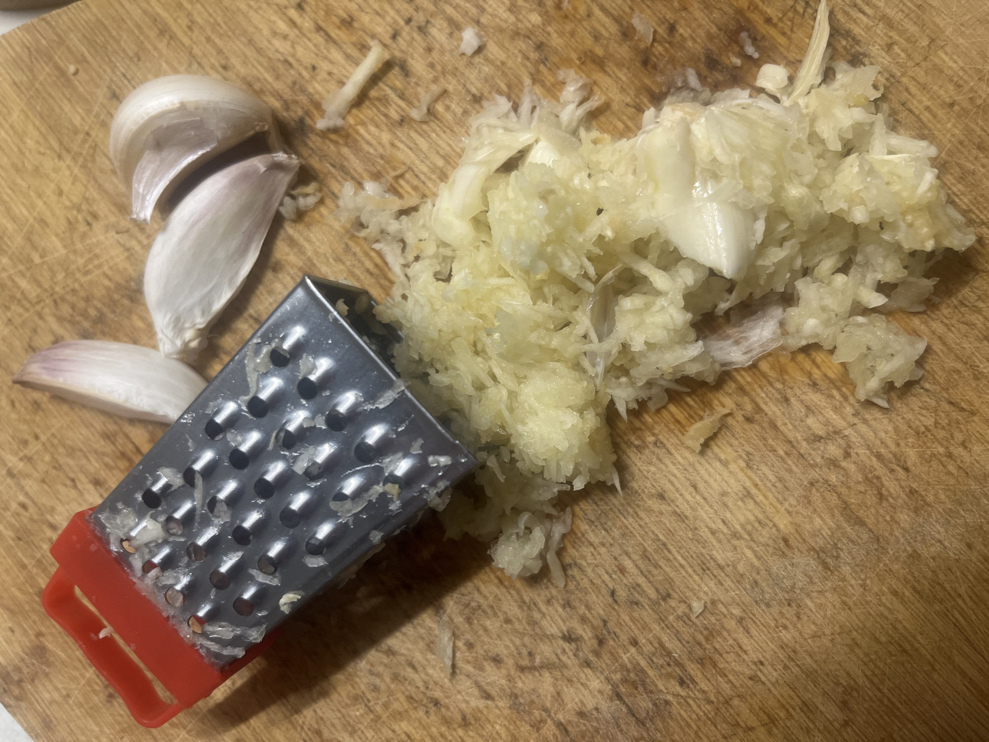 The image displays a close-up overhead view of a wooden cutting board with grated garlic and garlic cloves. A small, red and silver grater is positioned on the board, with finely minced garlic piled around it. Several unpeeled garlic cloves are also visible on the board, some appearing to be whole and others split open. The wooden surface of the cutting board shows signs of use, with visible grain and faint scratches. The lighting suggests an indoor setting, possibly a kitchen. There is no indication of the location being in Białystok, Poland, or any other specific city.