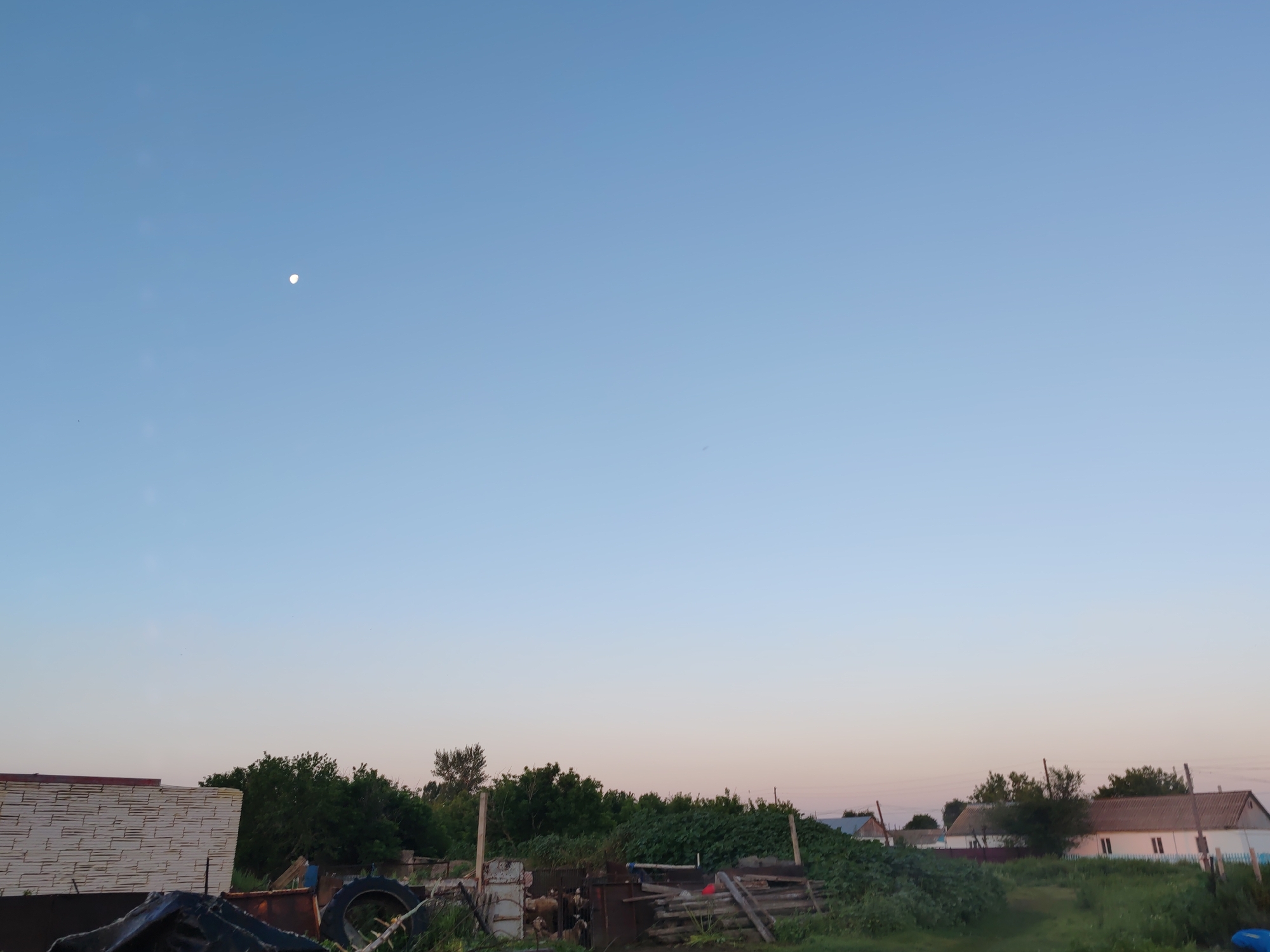 The image depicts a rural scene with a clear, pale blue sky, featuring a small crescent moon in the upper left quadrant. The horizon is lined with dense green foliage, interspersed with trees. In the foreground, there are various discarded objects, including large tires, metal sheeting, and wooden planks. To the left, a structure with horizontal white siding is partially visible. Further to the right, several houses with tiled roofs are situated. The overall impression is one of a developing or somewhat unkempt rural residential area, likely in the outskirts of Акжар, Kazakhstan, given the context provided. There are no people visible in the image.