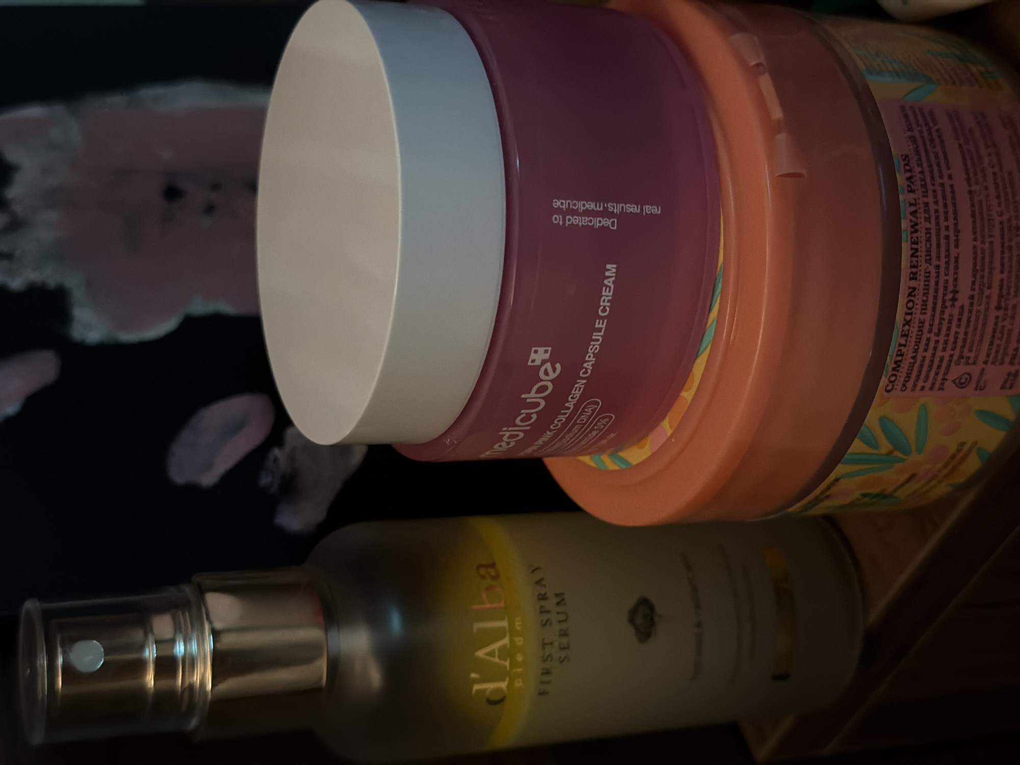 The image shows a close-up of several skincare products arranged on a surface, likely a shelf or countertop. On the left is a tall, clear bottle with a gold pump dispenser and a label that reads "d'Alba first spray serum." To the right of the serum is a round, pink jar with a white lid. The label on the pink jar is partially visible and appears to say "medicube pink collagen capsule cream." Below the pink jar is another round, orange jar with a decorative pattern on its label, which reads "COMPLEXION RENEWAL PADS." The background is dark and out of focus, with some blurred shapes that might be part of a poster or artwork. There are no people or animals in the image. The scene is static and does not depict any activity. The location in the city of Đà Nẵng, Vietnam, is not indicated by any visual cues in the image.