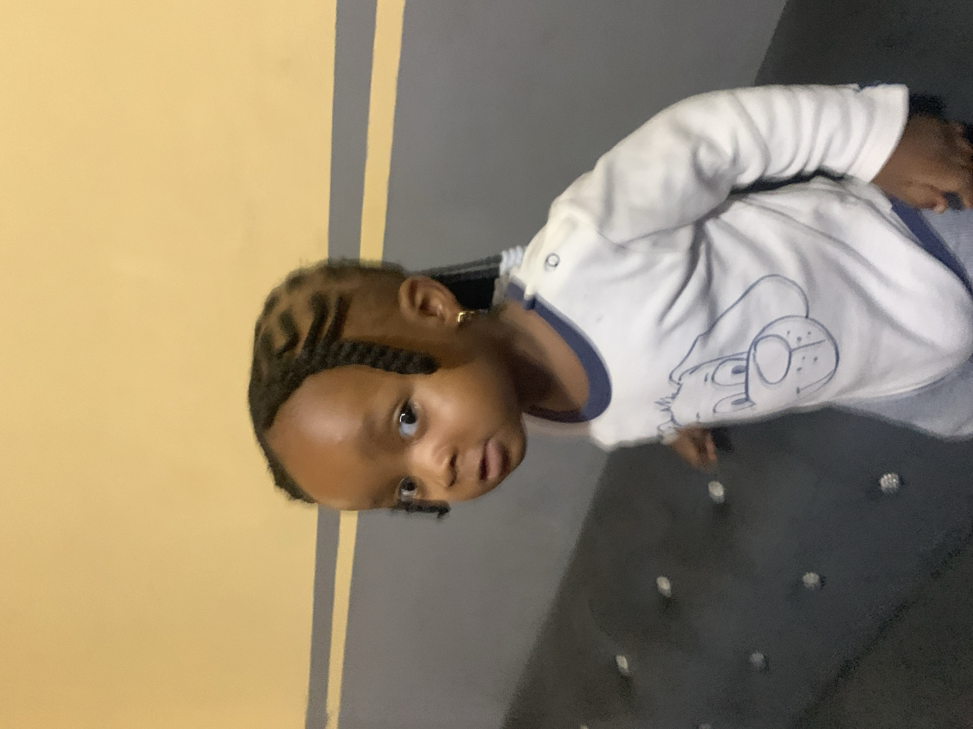 A young child with dark skin and braided hair is looking directly at the camera. The child is wearing a white long-sleeved shirt with a blue neckline and a cartoon dog design. The child has a gold hoop earring in their right ear. The background consists of a yellow and gray striped wall. The child is positioned in front of a dark, textured surface that appears to be upholstery, possibly a couch or bench. The lighting suggests an indoor setting. The image appears to have been taken in Jos, Nigeria, but there are no specific visual cues within the image to confirm this location.