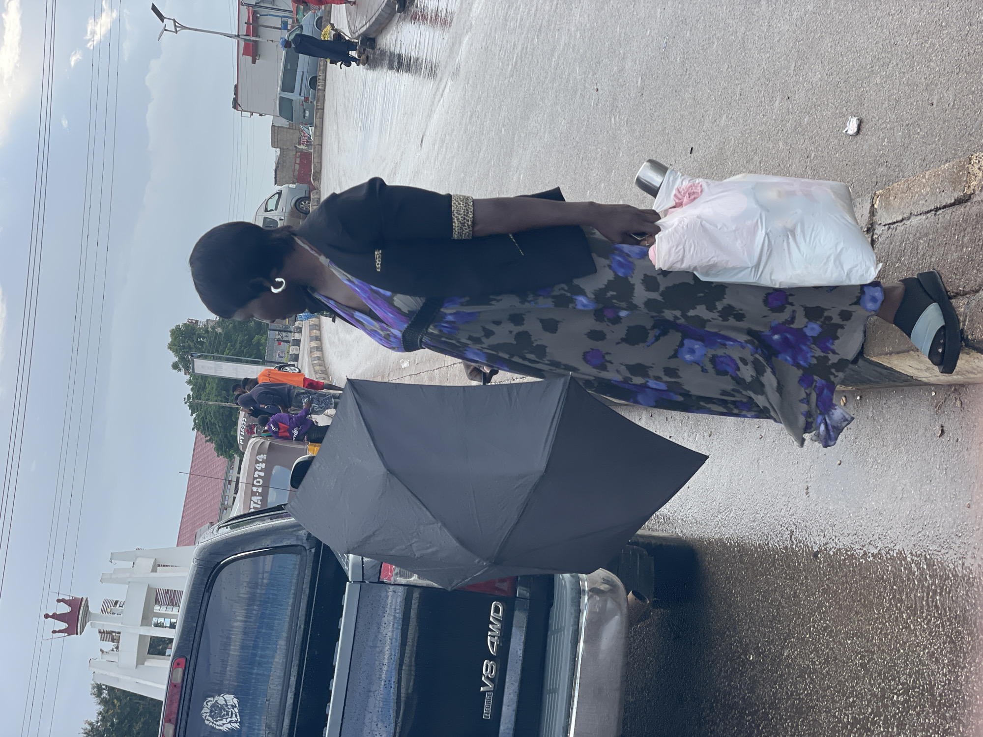 A woman stands near a black pickup truck on a wet street in Jos, Nigeria, holding a black umbrella and a white plastic bag. She wears a dark blazer over a floral print dress and a pair of blue and black sandals. In the background, a white monument with a red crown on top is visible, along with several vehicles and other people. The ground is wet, indicating recent rain. The scene appears to be an urban environment.