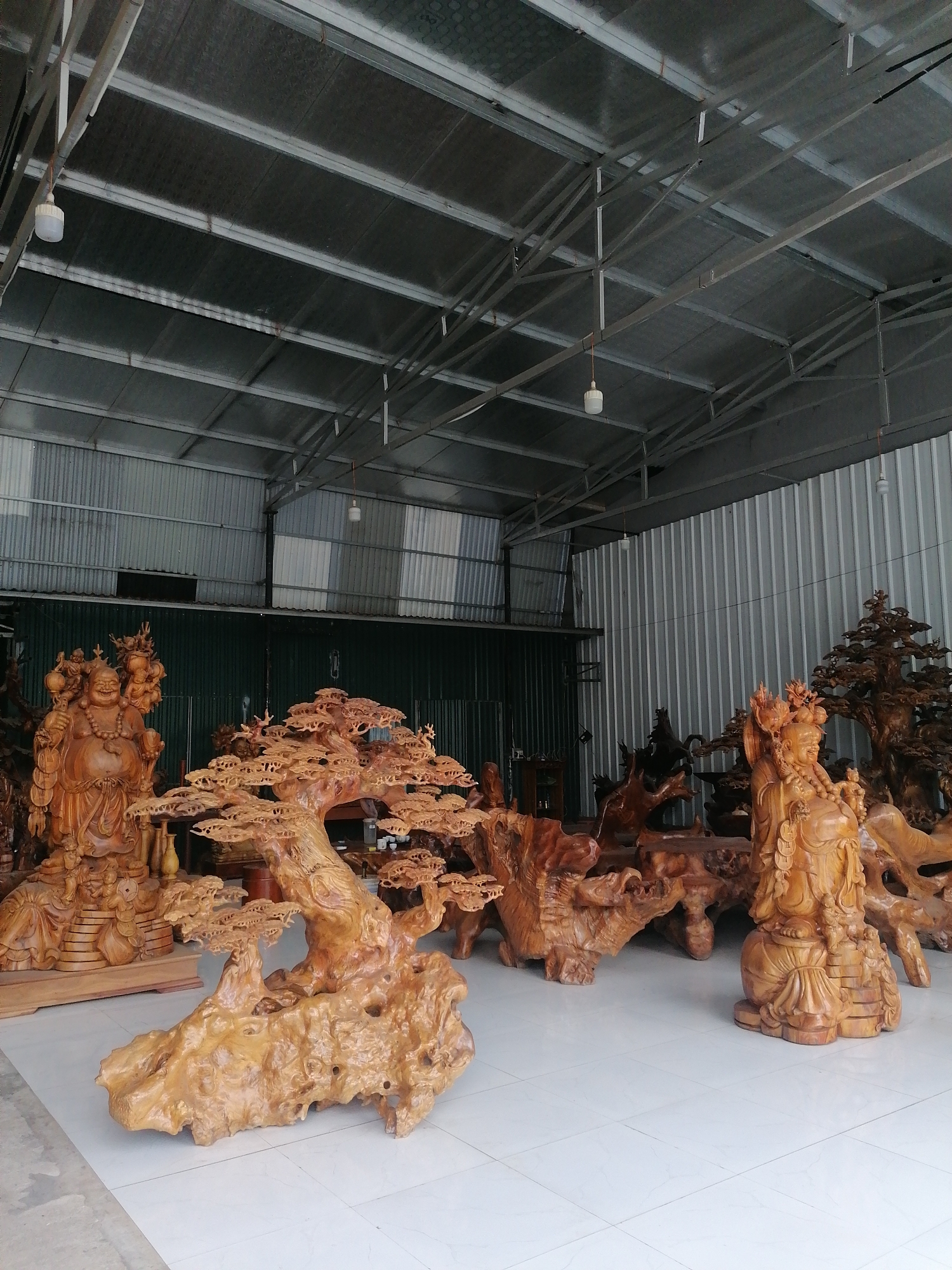 The image depicts a warehouse-like space in Ha Tu Ward, Vietnam, filled with wooden carvings. The space has a corrugated metal roof supported by a metal frame. Inside, numerous intricate wooden sculptures are displayed on a white tiled floor. Prominent among these are a carving of a laughing Buddha figure holding a sack, a tree-like sculpture with detailed foliage, and another standing figure. Other carved wooden items, possibly furniture or decorative pieces, are scattered throughout the room. The light in the warehouse is diffuse, suggesting natural light or fluorescent lighting. The background reveals more of the warehouse structure, including what appears to be a closed metal door.