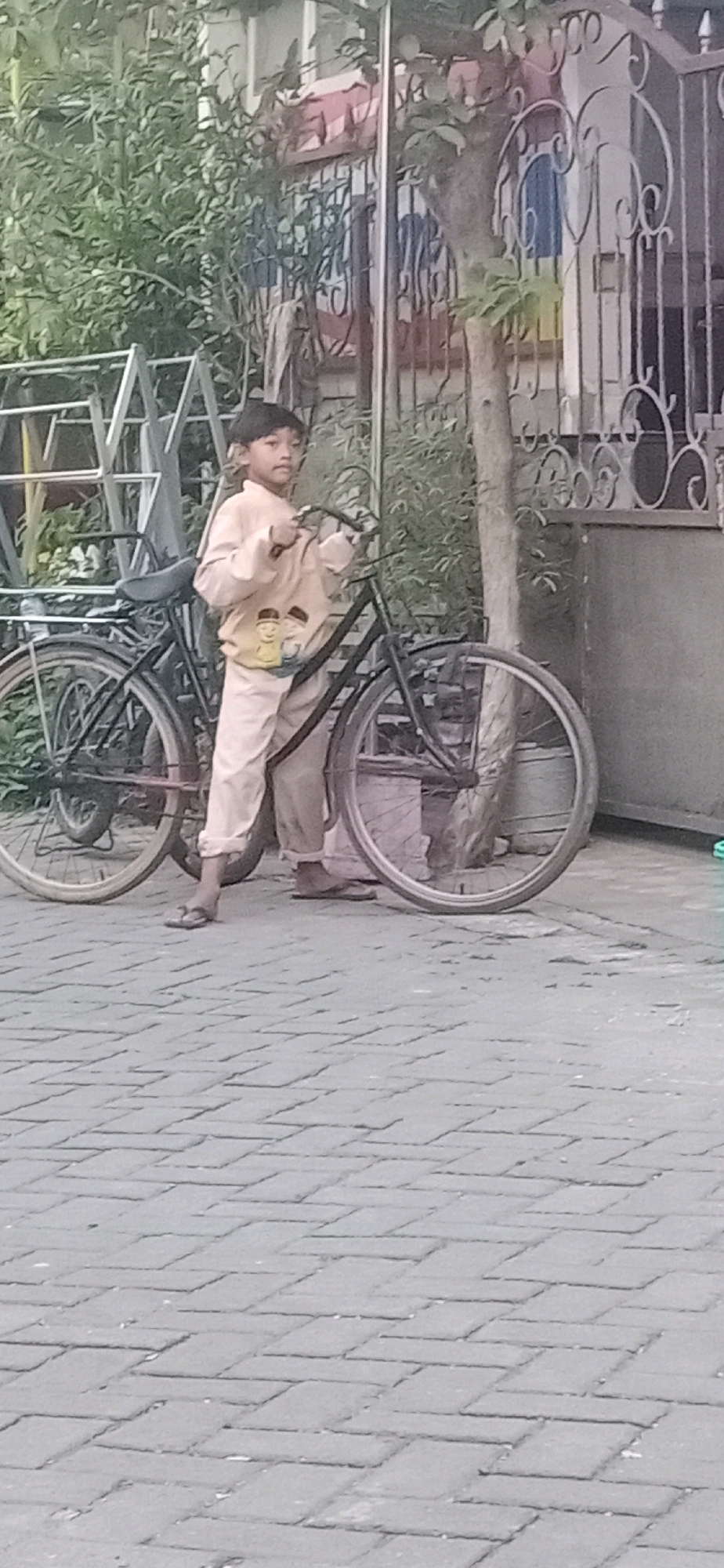 A young boy, appearing to be of East Asian descent, stands beside a bicycle on a brick-paved sidewalk. He is wearing a light-colored long-sleeved shirt with a graphic on the front depicting two cartoon characters and light-colored pants. The boy is holding the handlebars of the bicycle. The bicycle is dark-colored and appears to be an older model, with a front basket. Behind the boy and bicycle, there is dense green foliage and a portion of a building with an ornate metal fence. The background also shows a tree trunk. The scene appears to be an outdoor setting, possibly a residential area, in Sidojangkung, Indonesia.