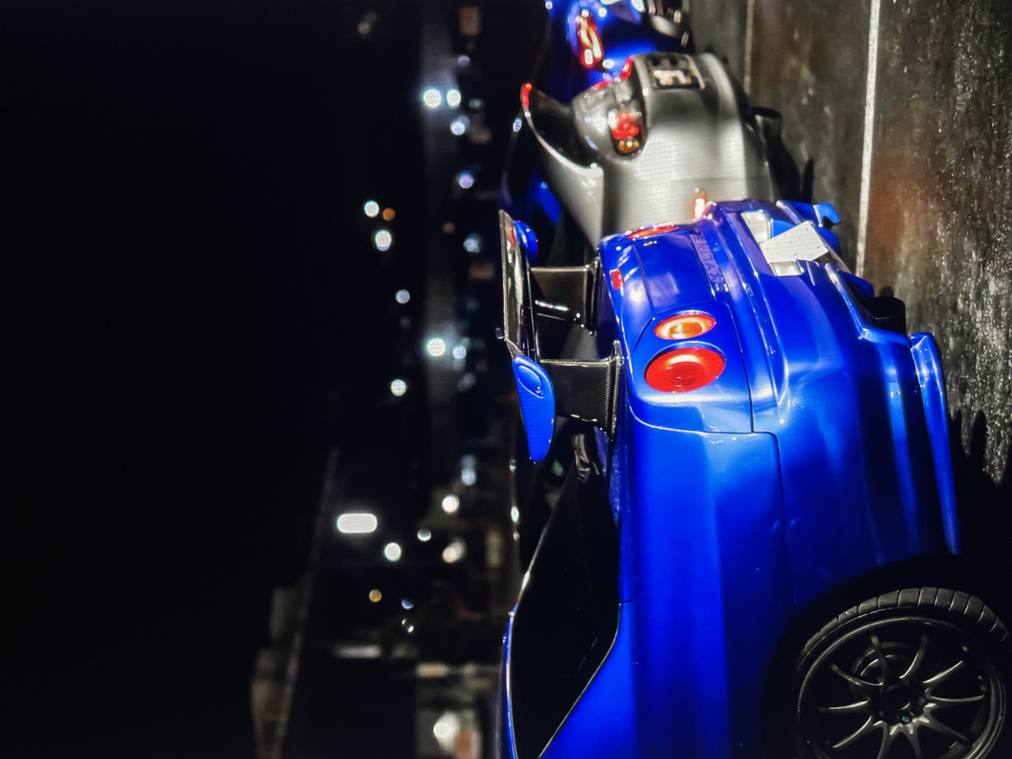 Here's a description of the image:
The image shows two toy cars in what appears to be a dimly lit environment. The dominant car is blue and positioned prominently in the foreground. It has a visible spoiler and taillights. Behind it, partially obscured, is a gray car. Bokeh lights are present in the background, suggesting an out-of-focus city scene. The scene implies an indoor location with an external, possibly urban view.